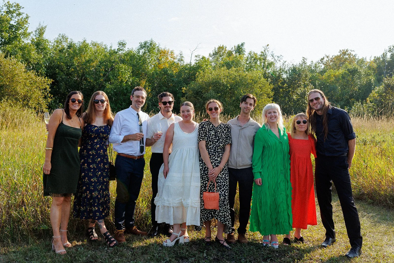A group of eleven people standing outdoors in a grassy field with trees in the background, dressed in semi-formal attire, some holding glasses of champagne, smiling at the camera.