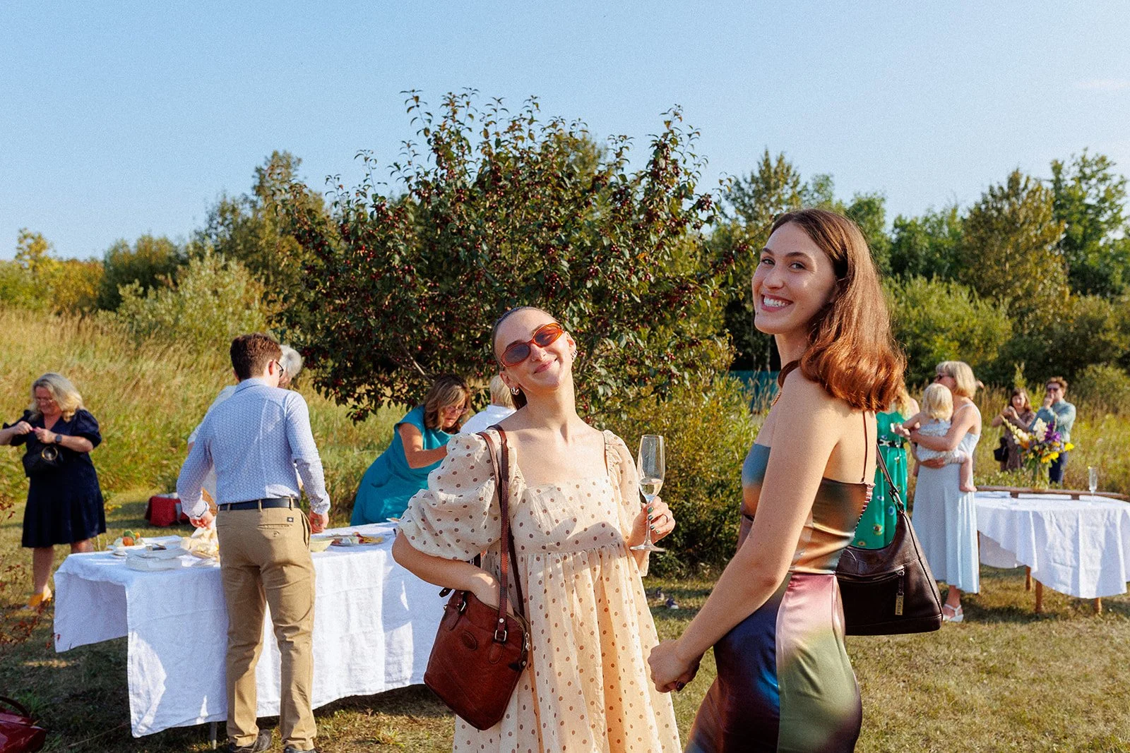 People at an outdoor gathering, with two women smiling at the camera in the foreground, one holding a glass of champagne, surrounded by others near tables with food and decorations on a bright, sunny day.