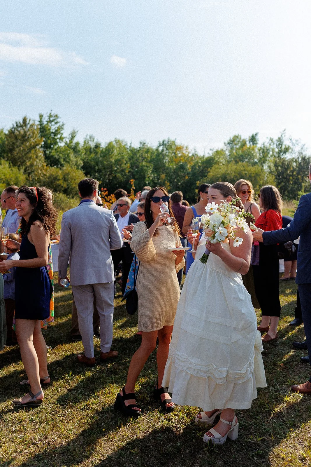 Group of people at an outdoor event, with a woman in a white dress holding a floral bouquet, and another woman in sunglasses drinking wine, on a grassy area with trees in the background under a sunny sky.