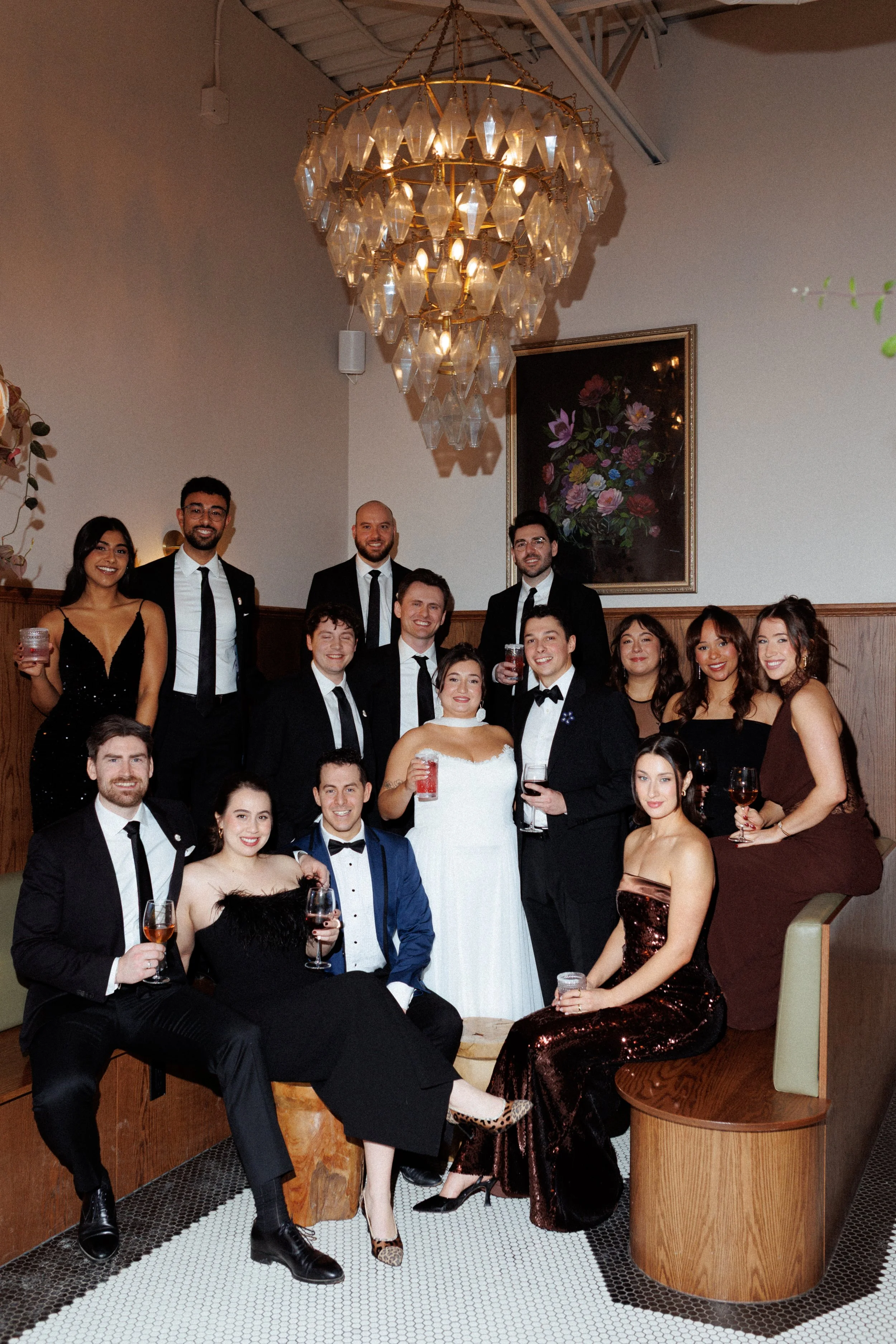 Group of people dressed in formal attire at a celebration or wedding, with a chandelier overhead and a floral painting on the wall.