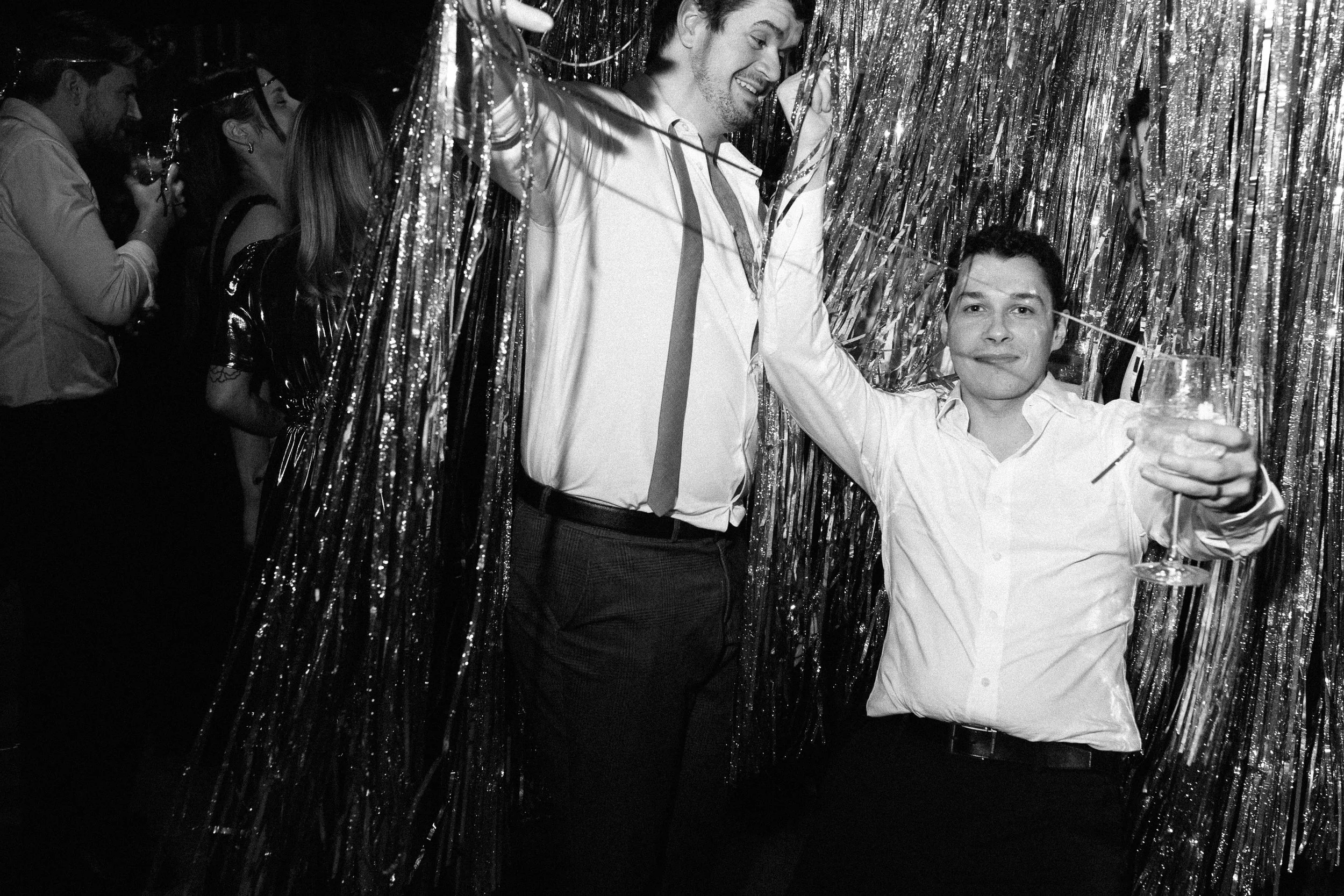 Two men celebrating at a party with metallic streamers in the background. One man is holding a glass of drink and a straw, and the other is smiling with his eyes closed.