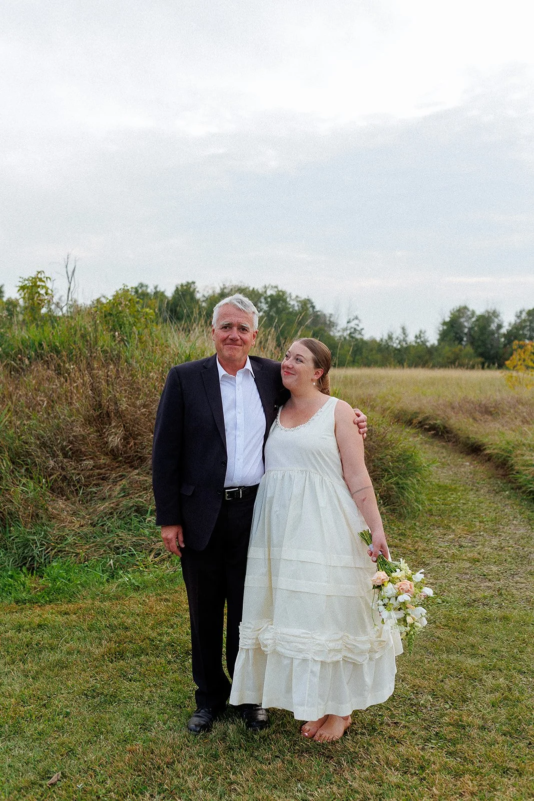 A bride in a white dress and a man in a suit standing outdoors on a grassy field with trees in the background.