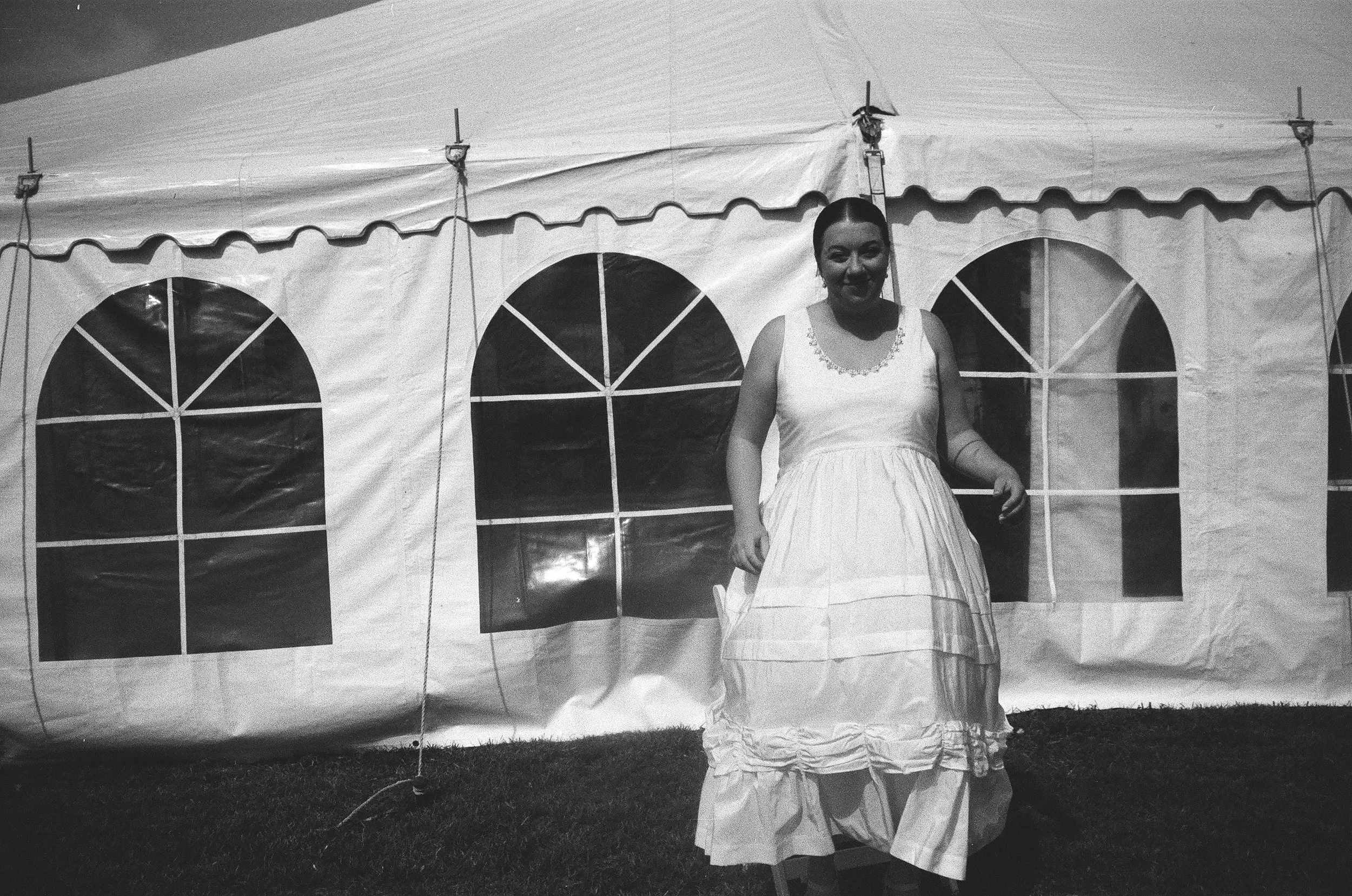 A woman dressed in a long, white sundress standing in front of a large tent with arched windows.