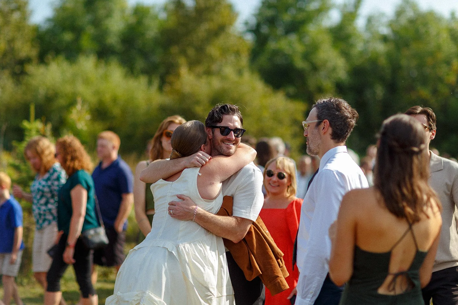 People embracing and socializing outdoors during a sunny day with trees in the background.
