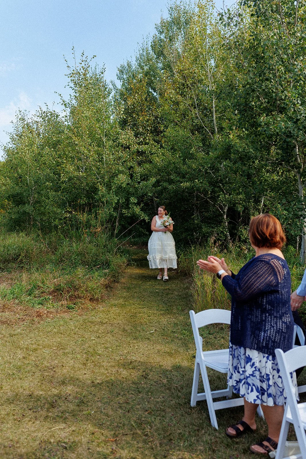 A bride in a white dress holding a bouquet walking down an outdoor aisle, with guests seated on white chairs clapping, surrounded by green trees and grass on a clear day.