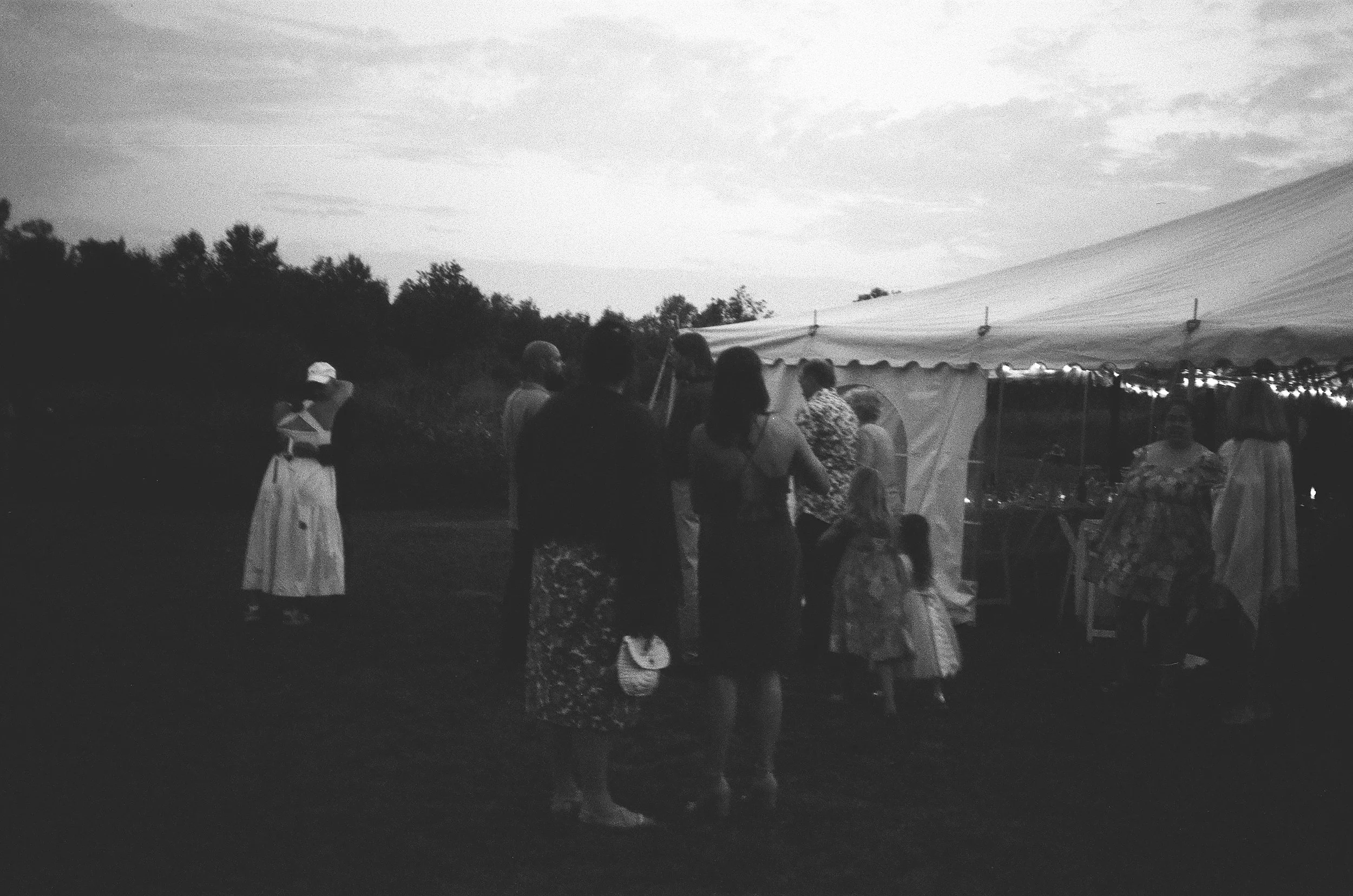 People gather near a large event tent outdoors in the evening, some standing and talking, others seated, with a tree line in the background.
