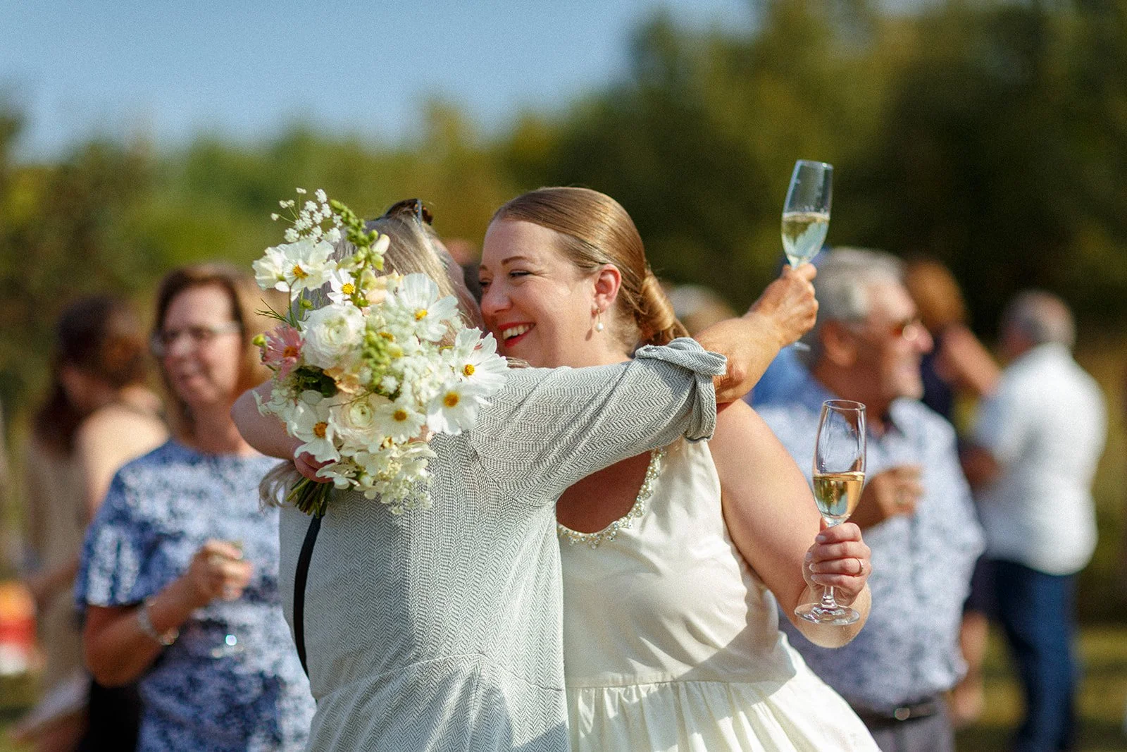 Two women hugging at a wedding reception, one holding a bouquet and the other holding a glass of champagne, with other guests in the background.