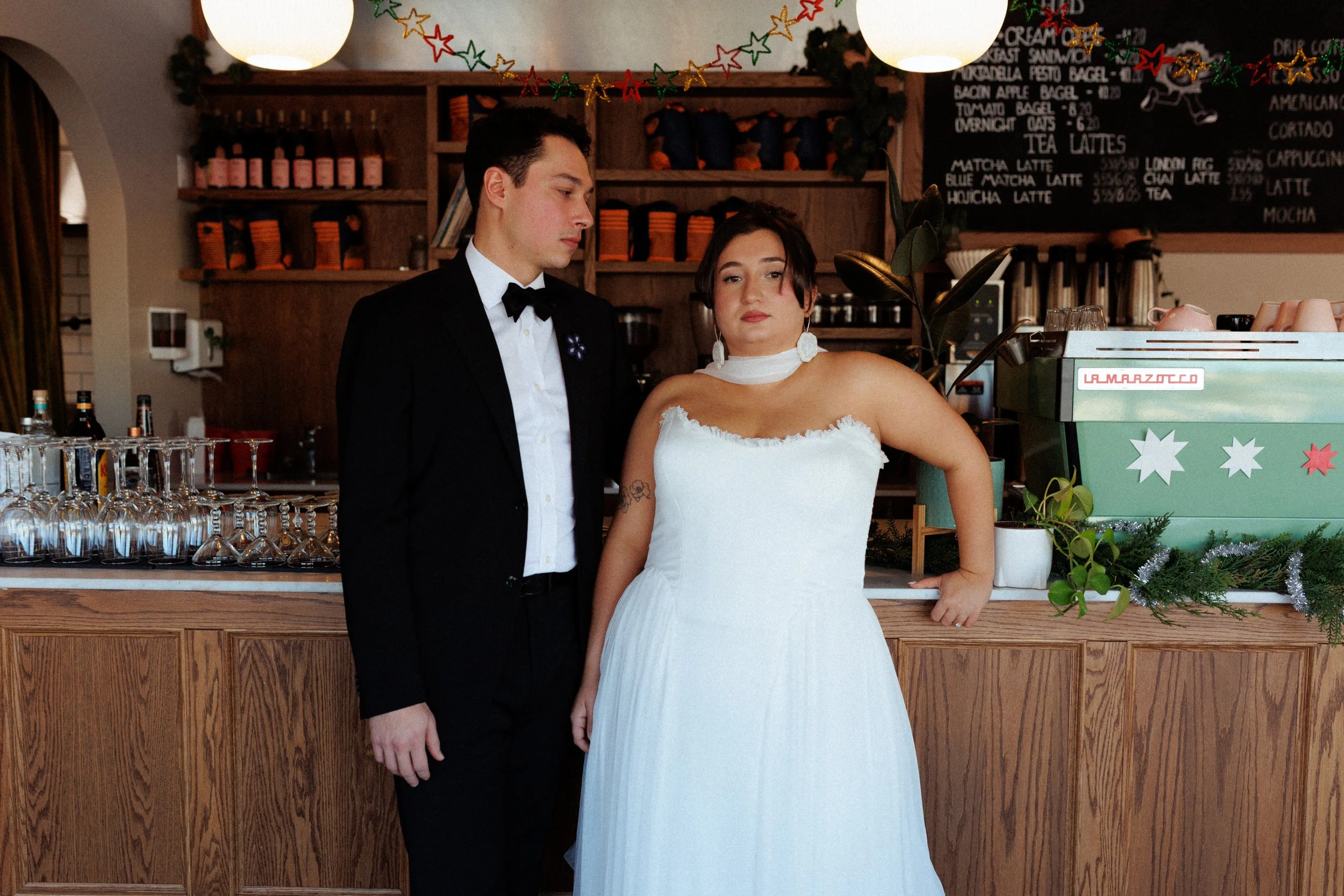 A young man in a black tuxedo and a young woman in a white strapless dress standing at a cafe counter, with a chalkboard menu behind them and a festive Christmas garland overhead.