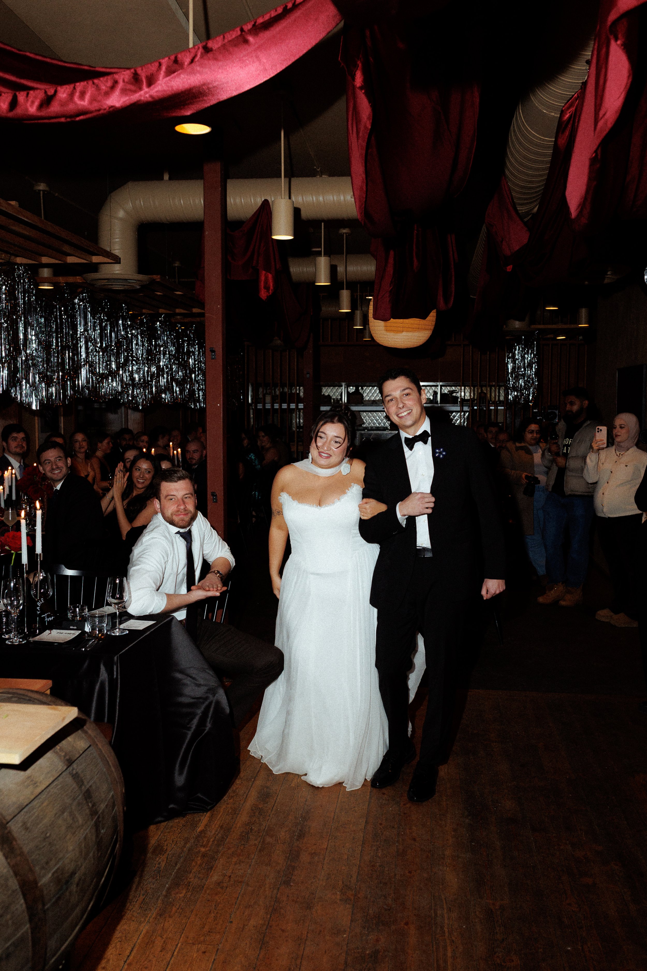 A bride and groom walking arm in arm down the aisle at their wedding reception, surrounded by seated guests and onlookers.