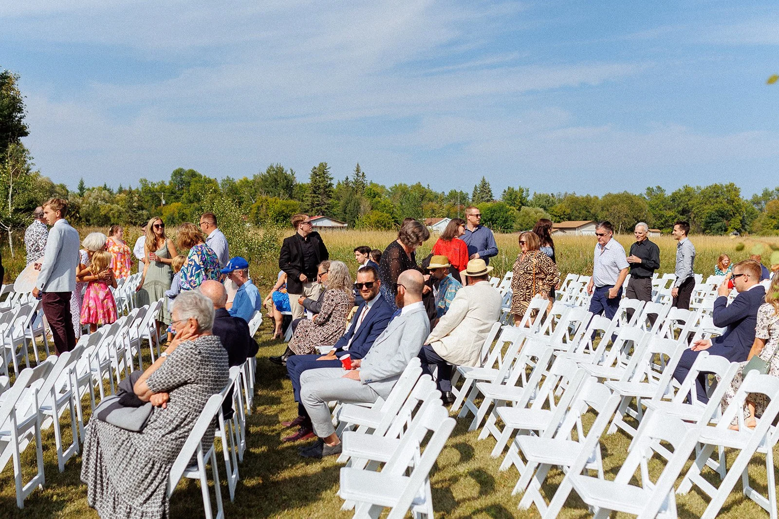 People attending an outdoor wedding ceremony during daytime, seated on white folding chairs in a grassy field with trees and houses in the background.