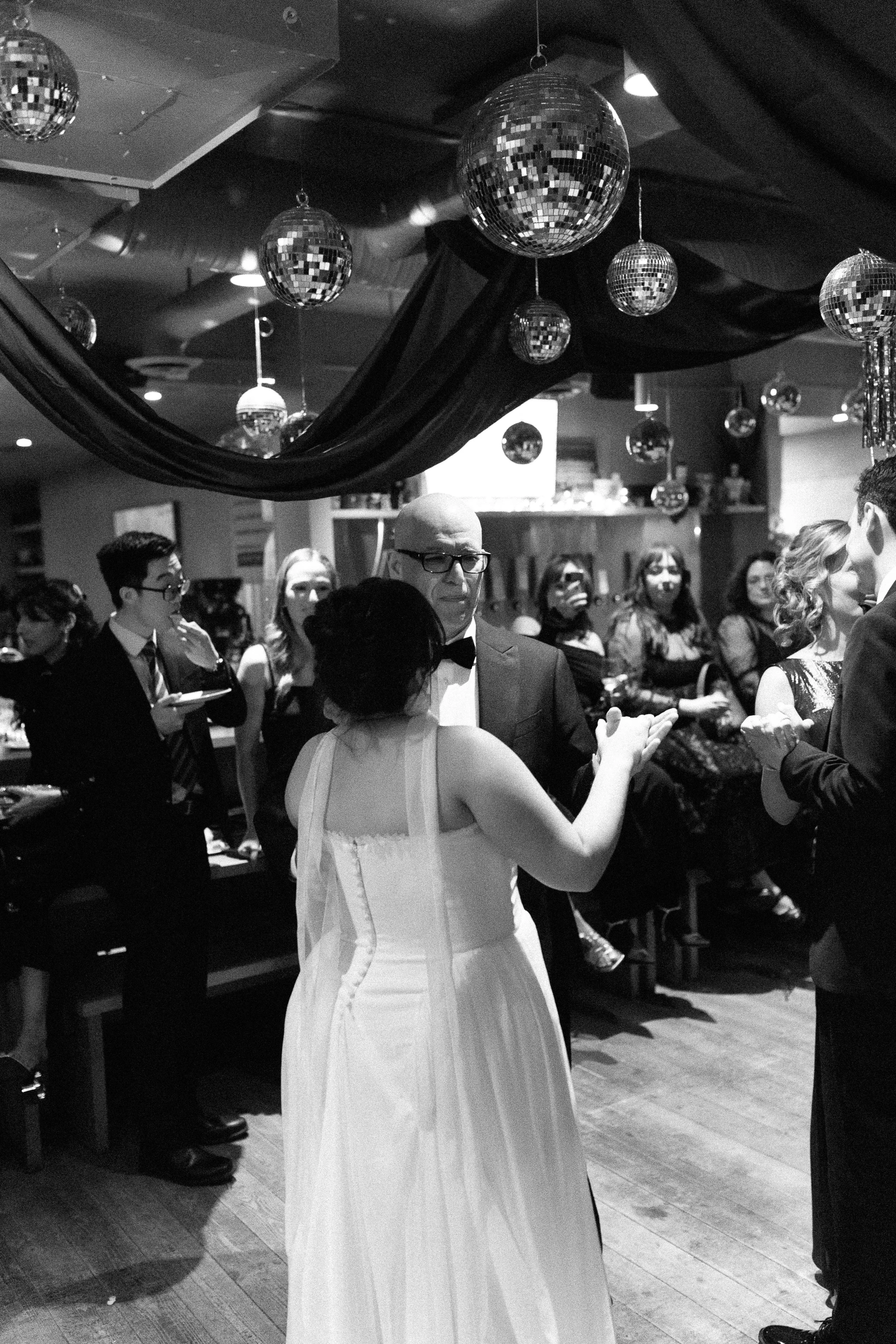 A black and white photo of a formal dance or gathering with couples dancing. The room is decorated with multiple disco balls hanging from the ceiling, and there is a large draped fabric.