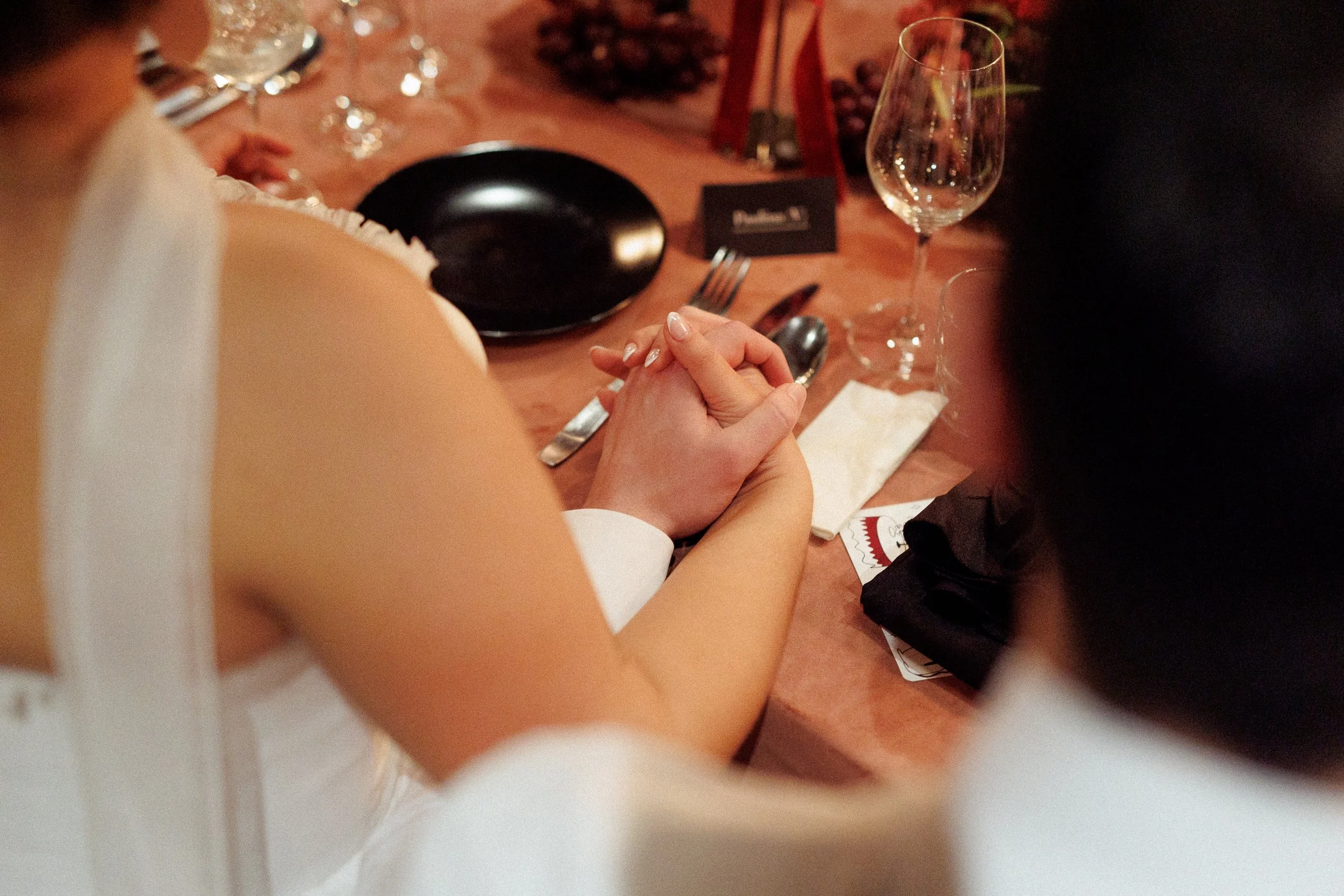 Close-up of a couple holding hands across a table at a formal event, with a black plate, a wine glass, silverware, and a name card on the table.