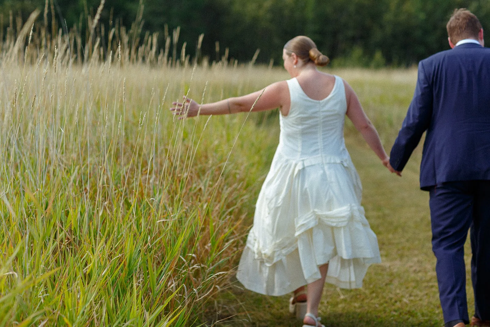 A woman in a white wedding dress and a man in a dark suit holding hands, walking through a grassy field.