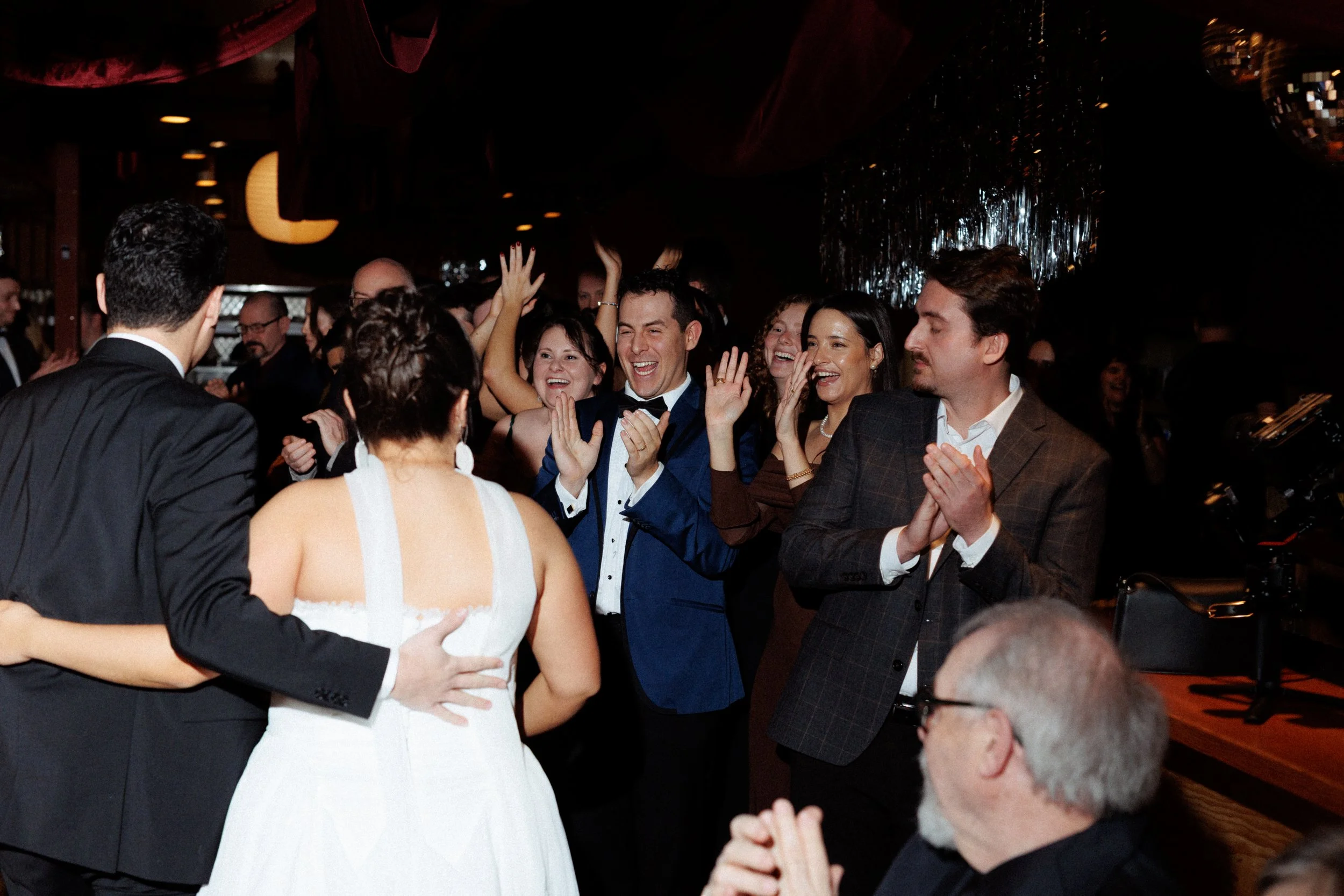 People celebrating at a wedding reception, smiling and clapping, with a bride and groom in the foreground and guests in the background.