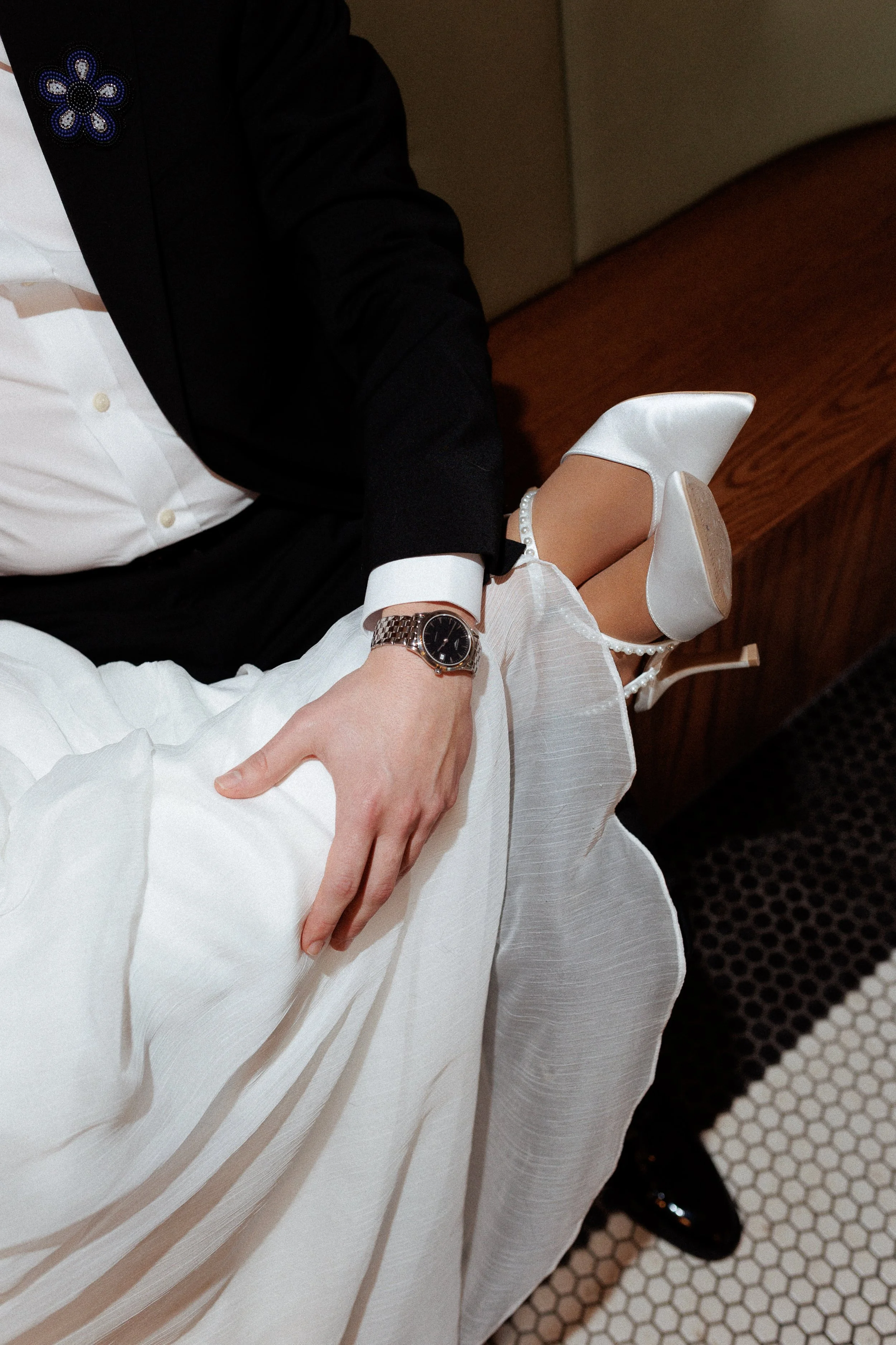 A person dressed in formal attire, including a black jacket, white shirt, and jewelry, seated on a bed with a white dress, white high heels, and a watch on their wrist.