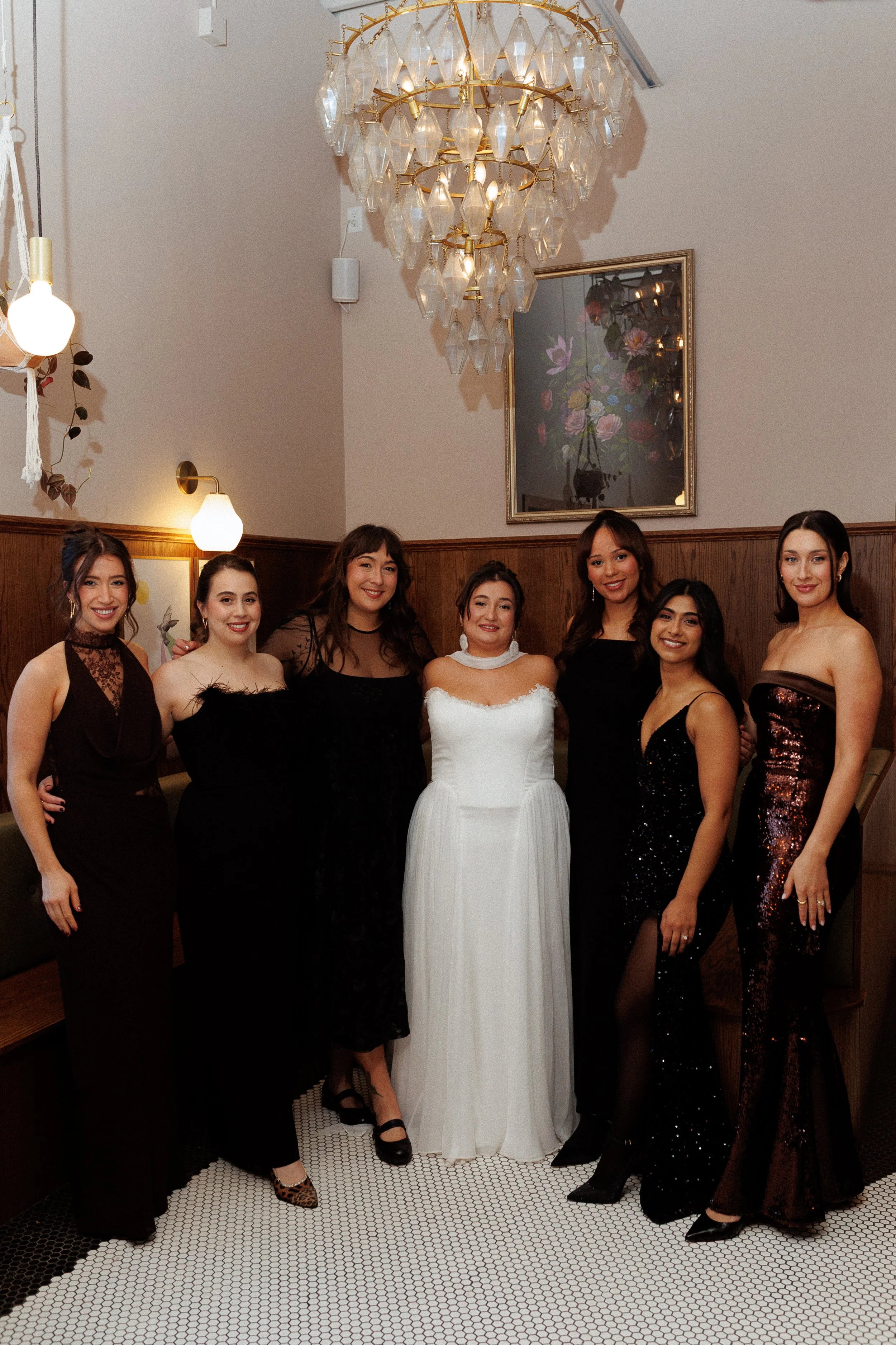 Seven women dressed in elegant evening gowns standing together in a warmly lit room with a chandelier, wooden paneling, and framed artwork on the wall.