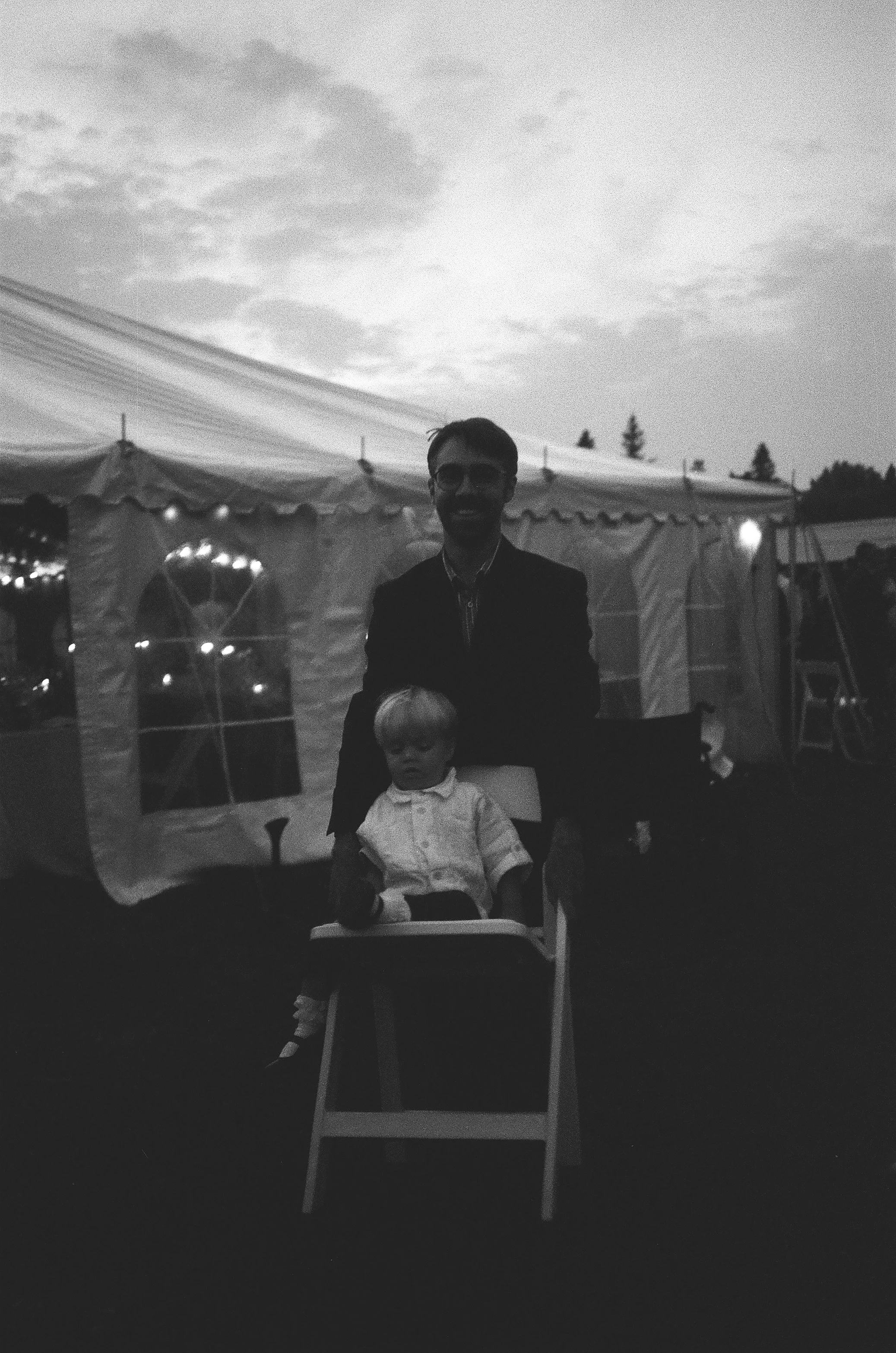 Black and white photo of a man and a young boy inside a large outdoor tent, with the man standing behind the boy who is seated in a high chair. The background shows some trees and sky with clouds.