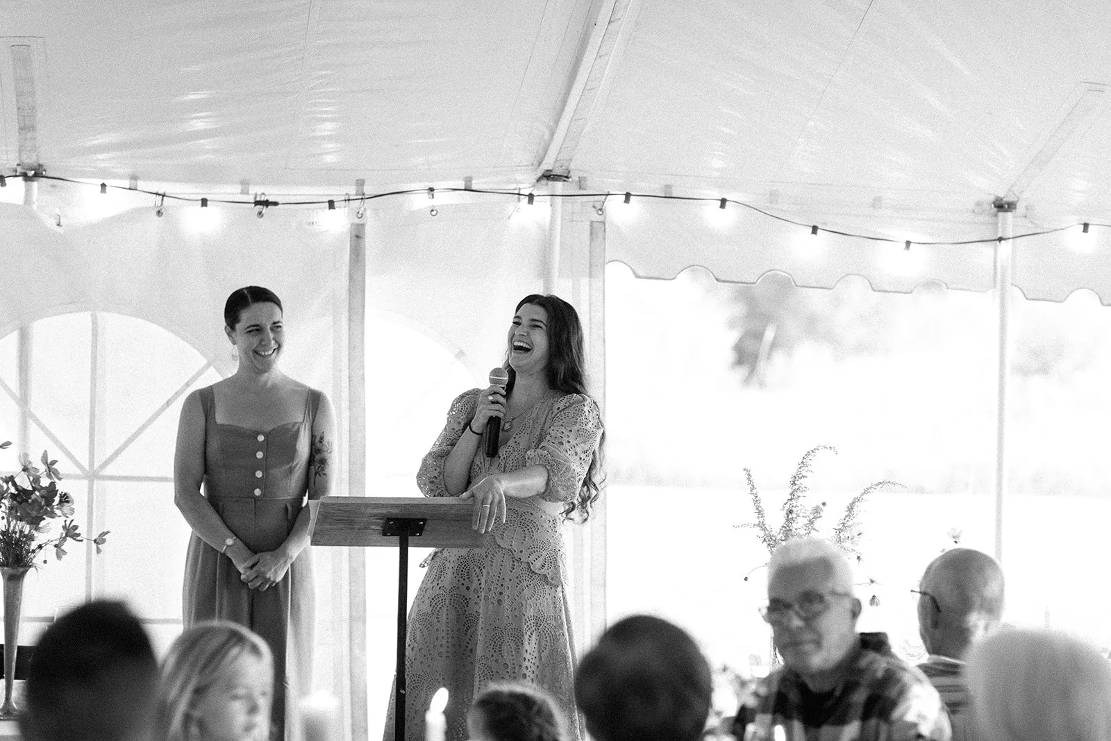Two women giving a speech at a gathering inside a tent, with people seated at tables in the foreground, one woman holding a microphone and laughing, the other smiling.