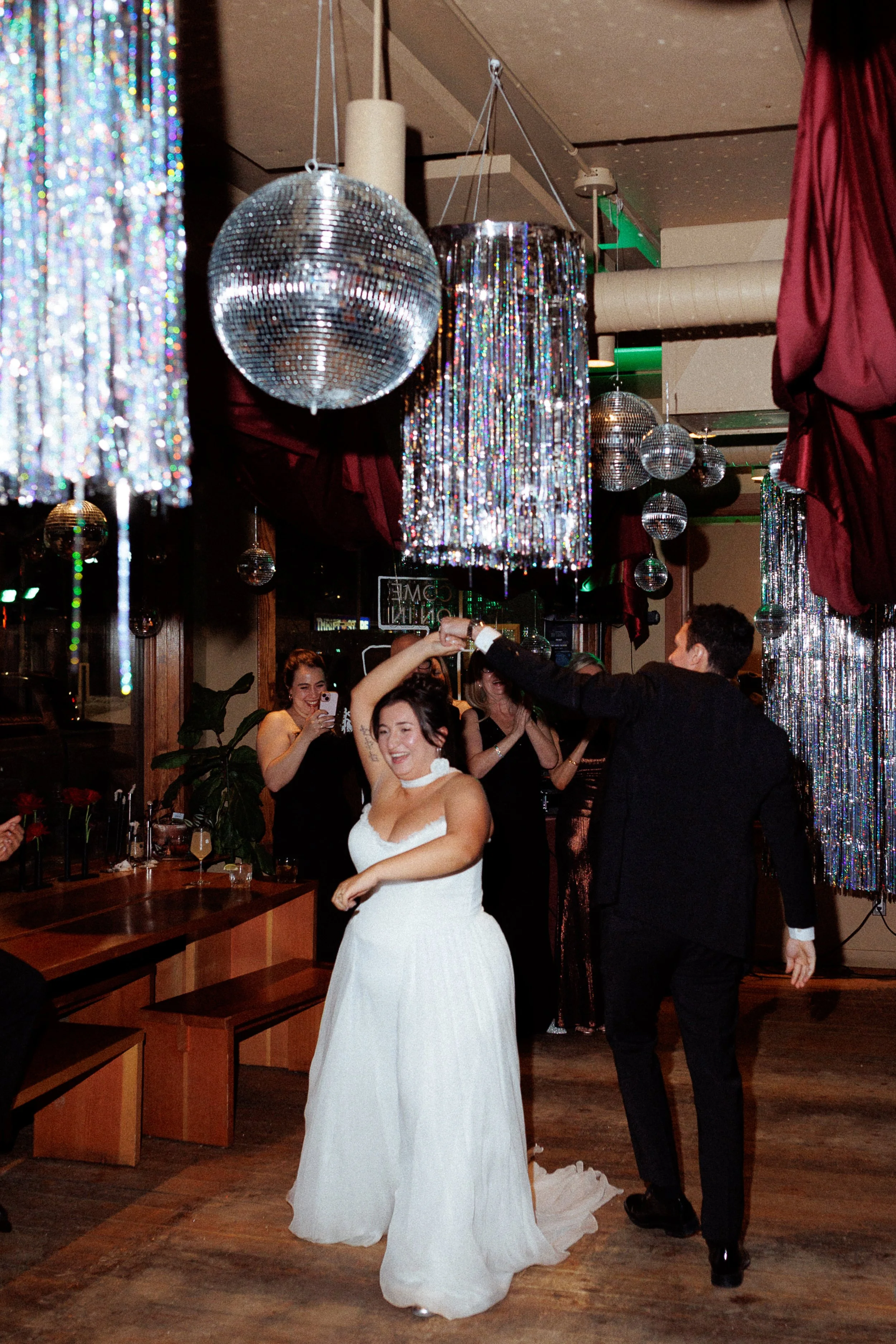 A bride in a white wedding dress and a groom in a black suit dance together at a celebration, surrounded by party decorations with hanging disco balls and shiny curtains, with guests smiling and taking photos.