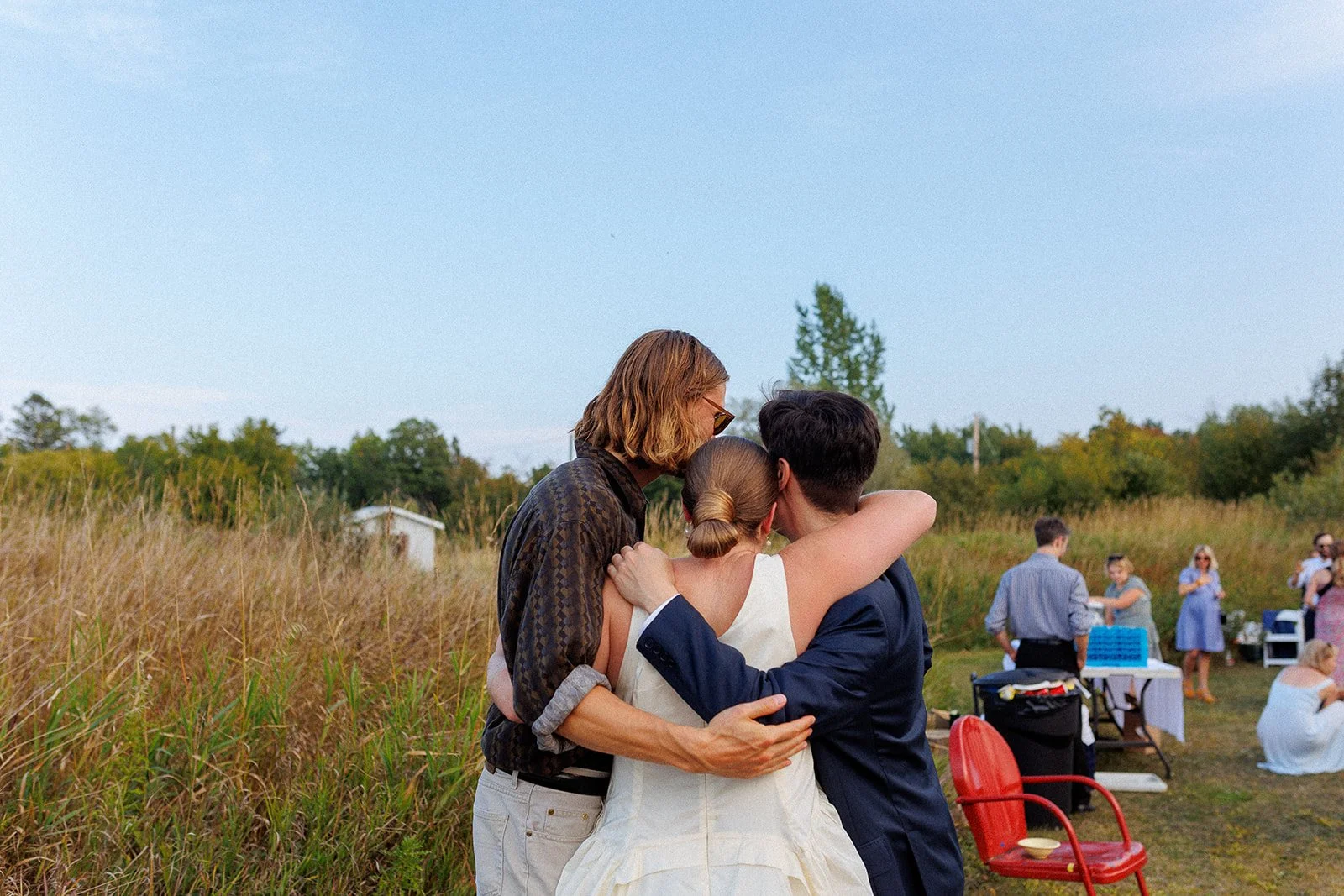 Three people embrace during an outdoor gathering, with others in the background and a grassy field and trees under a clear sky.