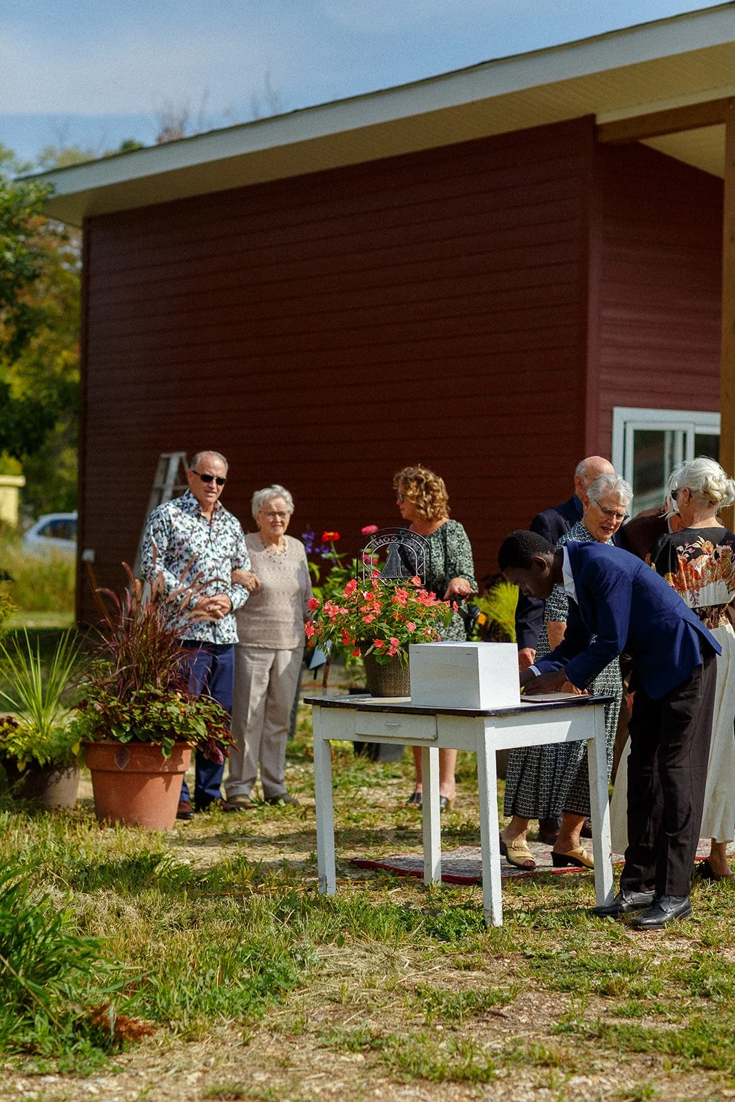Group of people outdoors gathered around a table with a flower pot and a white box, participating in a voting or signing activity, near a red building with a window.