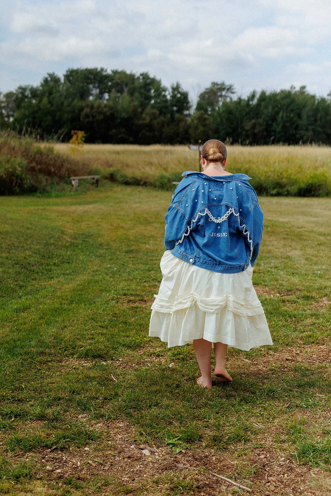 A person walking barefoot on a grassy path, wearing a white dress and a blue denim jacket with pearl decorations and the name 'Jesse' on the back, in an open outdoor field with trees in the background.