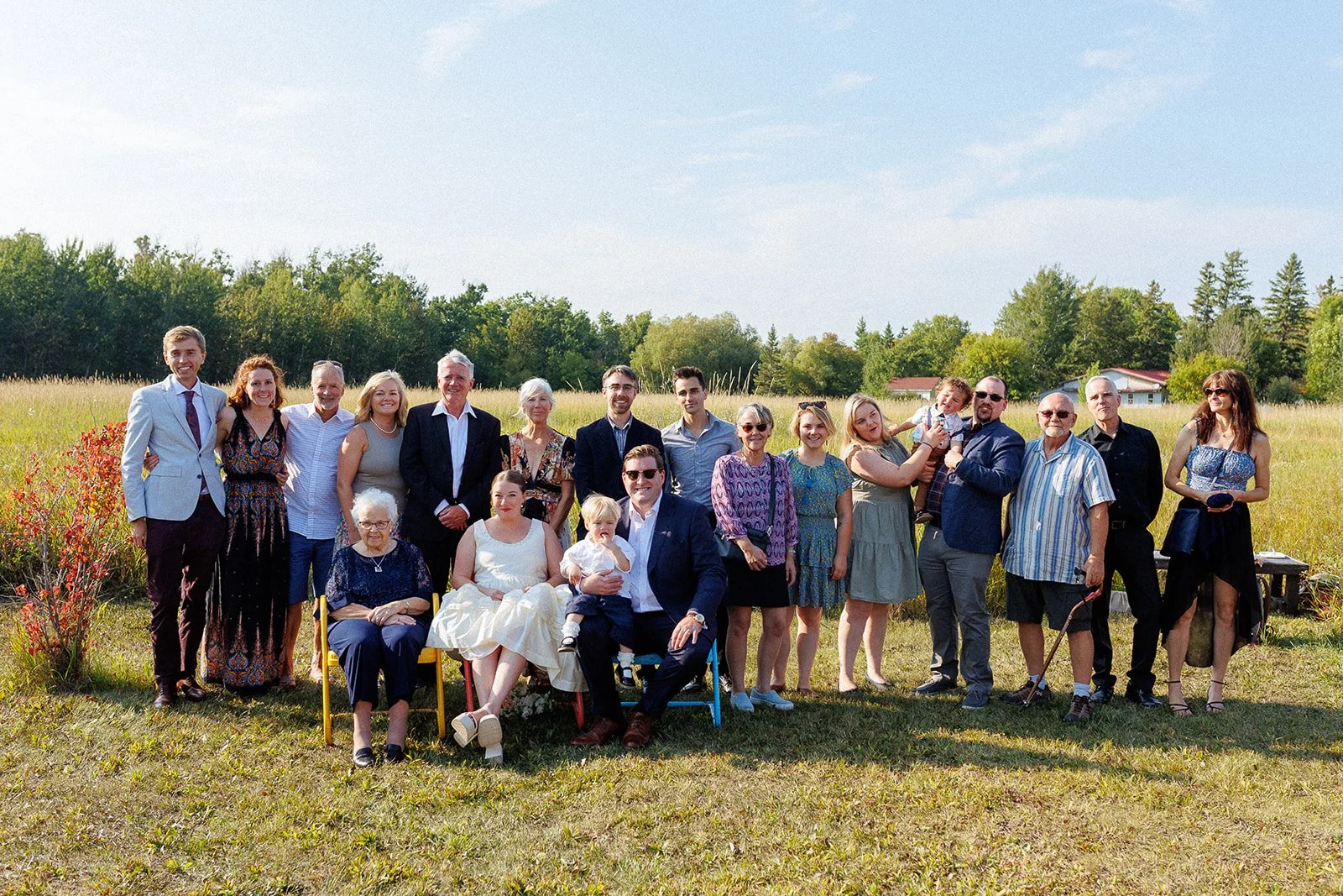 A large group of people posing outdoors in a grassy field, some sitting and some standing, under a partly cloudy sky with trees and small houses in the background.