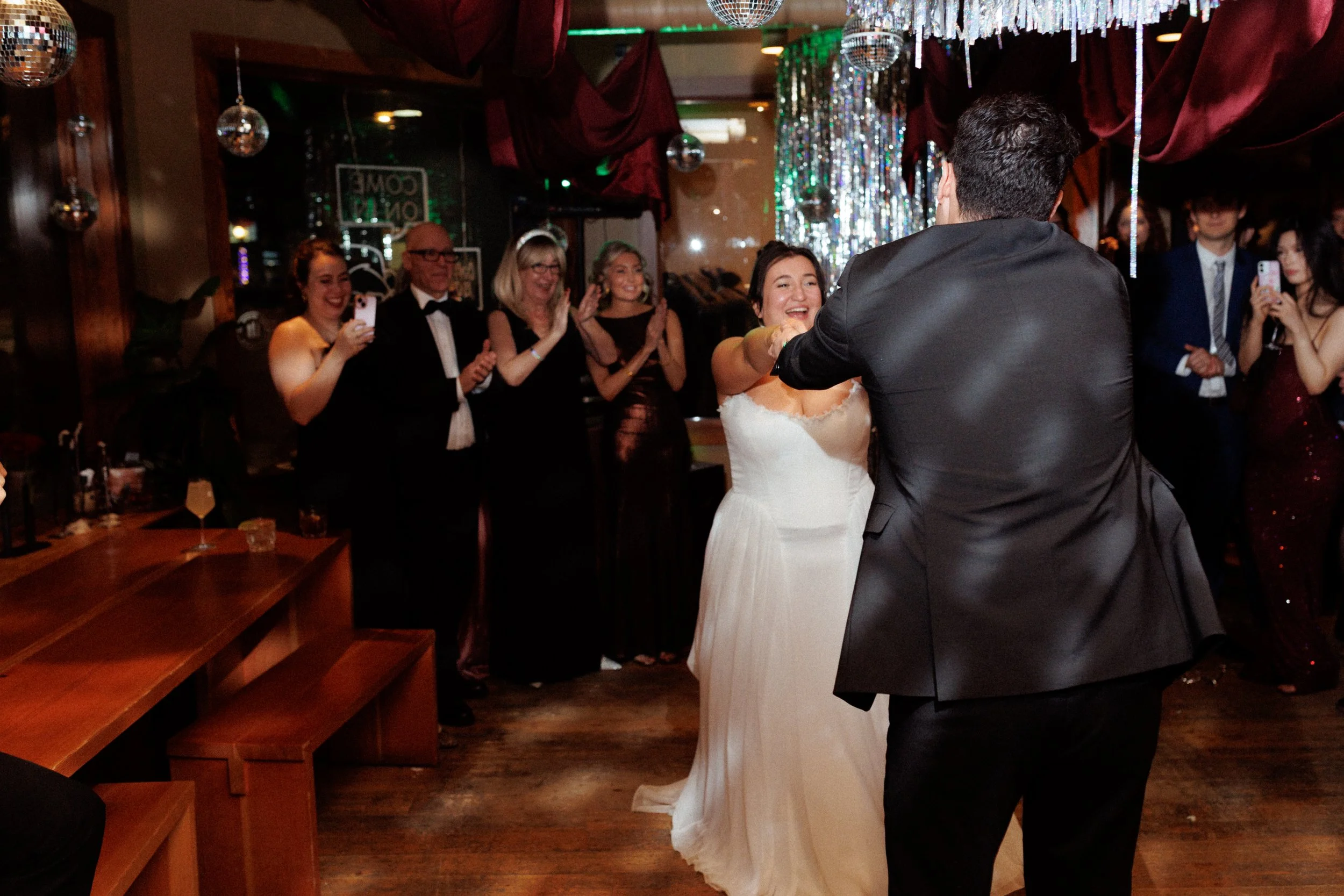 A bride and groom dance at their wedding reception as guests look on and cheer. The bride is smiling and reaching out to the groom, who is facing away from the camera. The guests are clapping and taking photos in a decorated (festive and elegant) ven