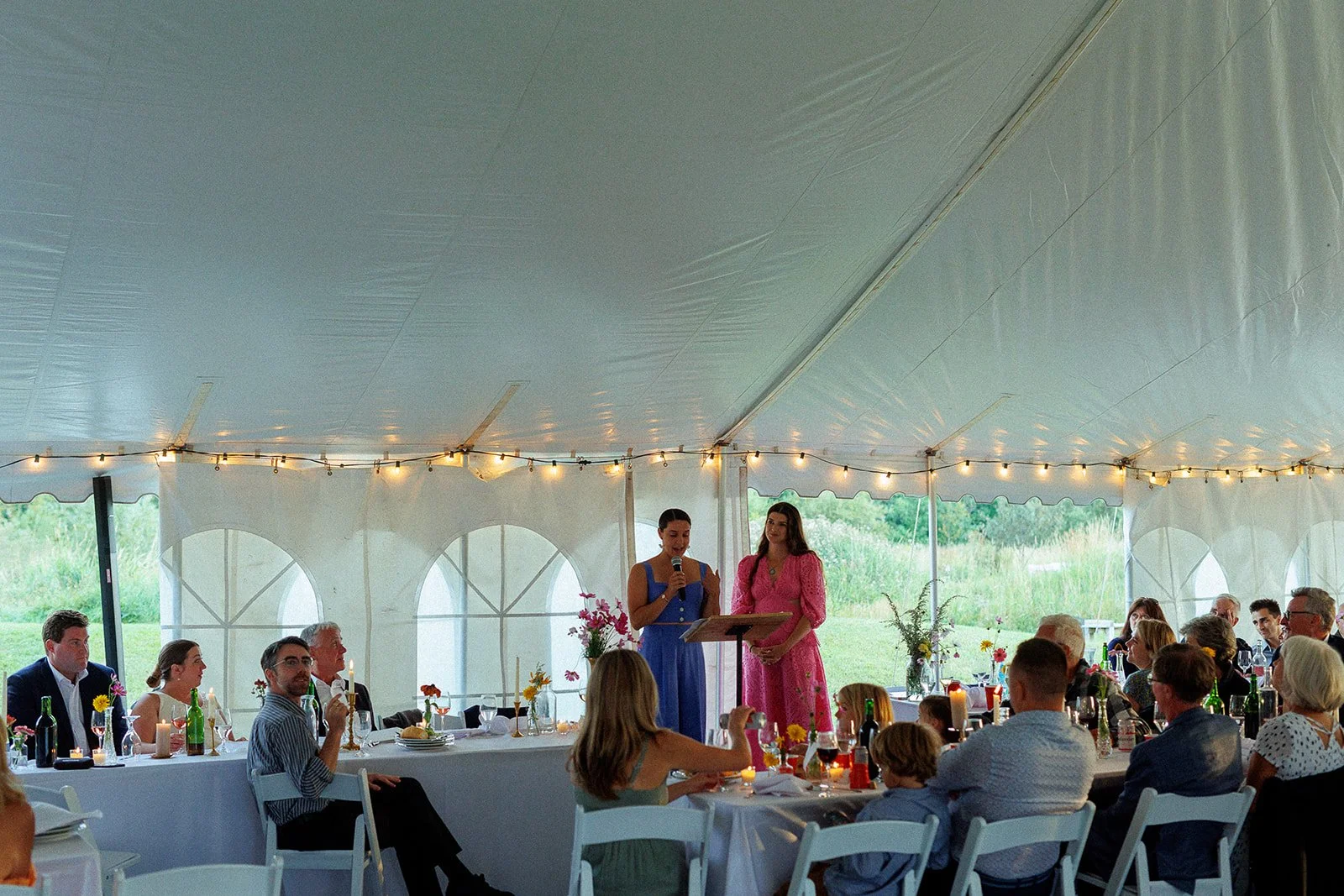 A wedding reception inside a large white tent with string lights, where two women are standing at the front, one speaking into a microphone and the other listening. Guests are seated at tables decorated with flowers and candles, with a lush green out
