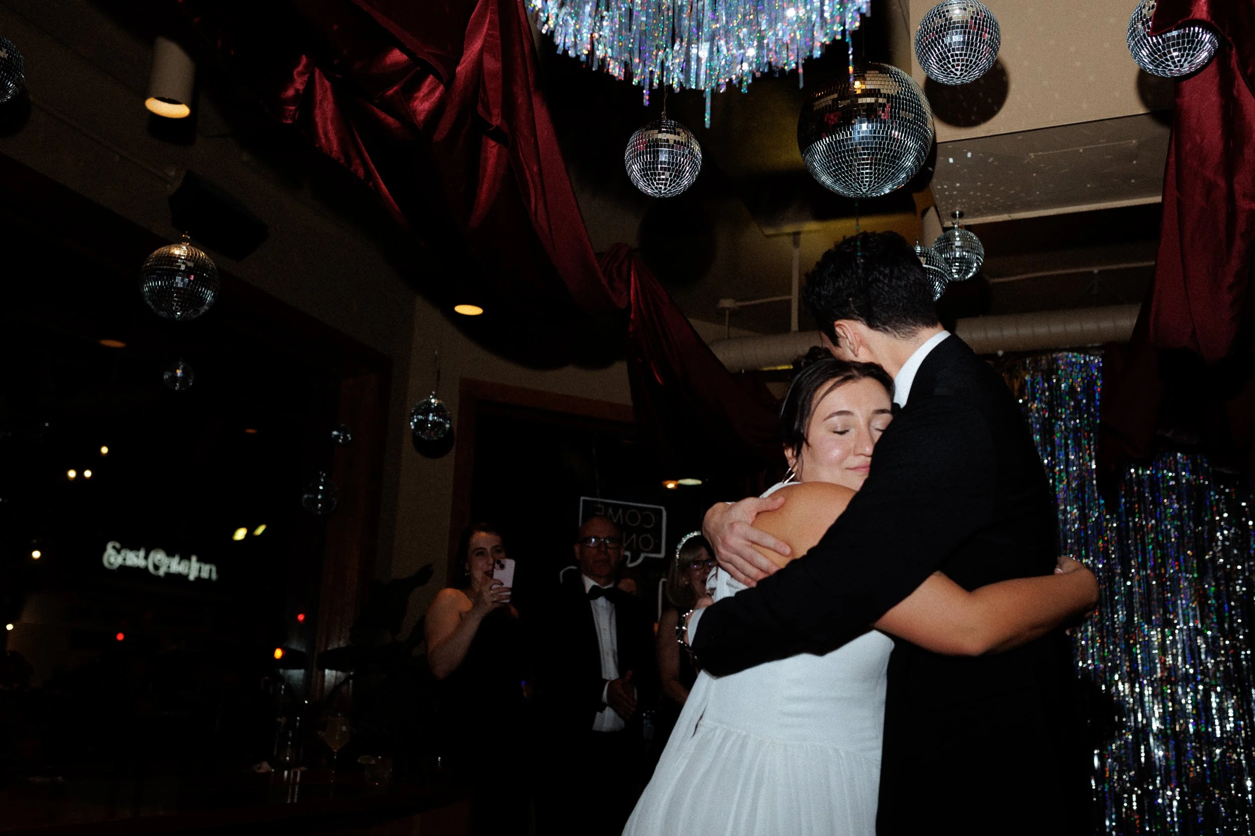 A couple sharing a dance at a wedding reception, with a woman in a white dress and a man in a black suit embracing. Disco balls and colorful decorations hang from the ceiling, and other guests are watching and taking photos in the background.
