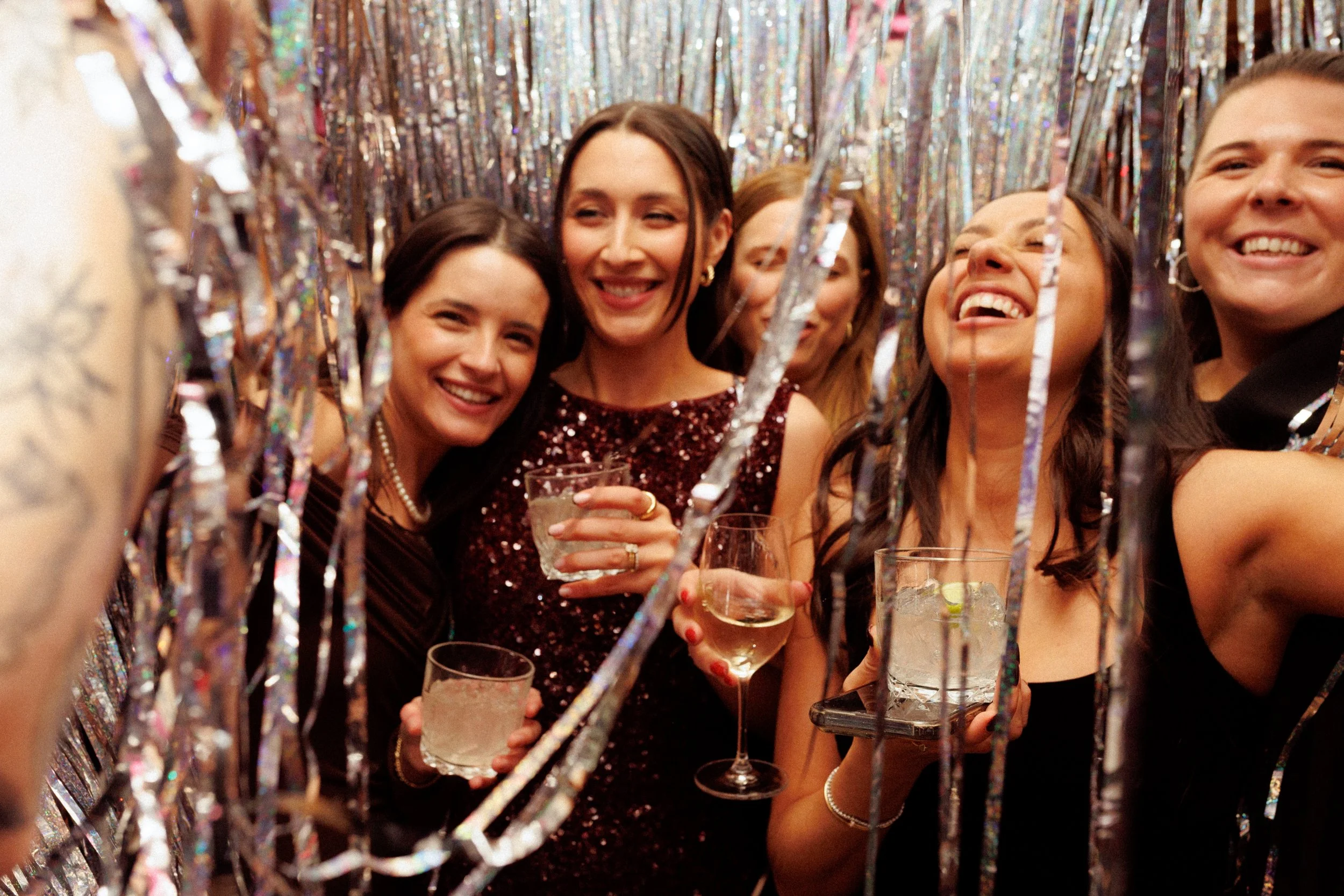 Group of women celebrating at a party with shiny decorations, wearing elegant dresses, holding drinks, and smiling.