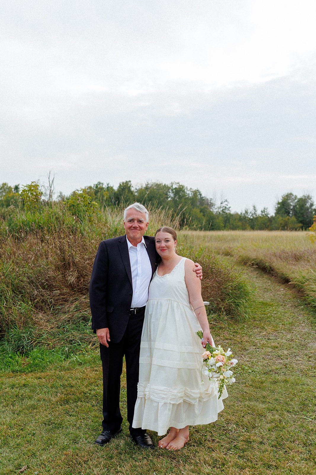 A man in a black suit and a woman in a white dress standing together outdoors on a grassy field, holding a bouquet of flowers, with trees and a cloudy sky in the background.