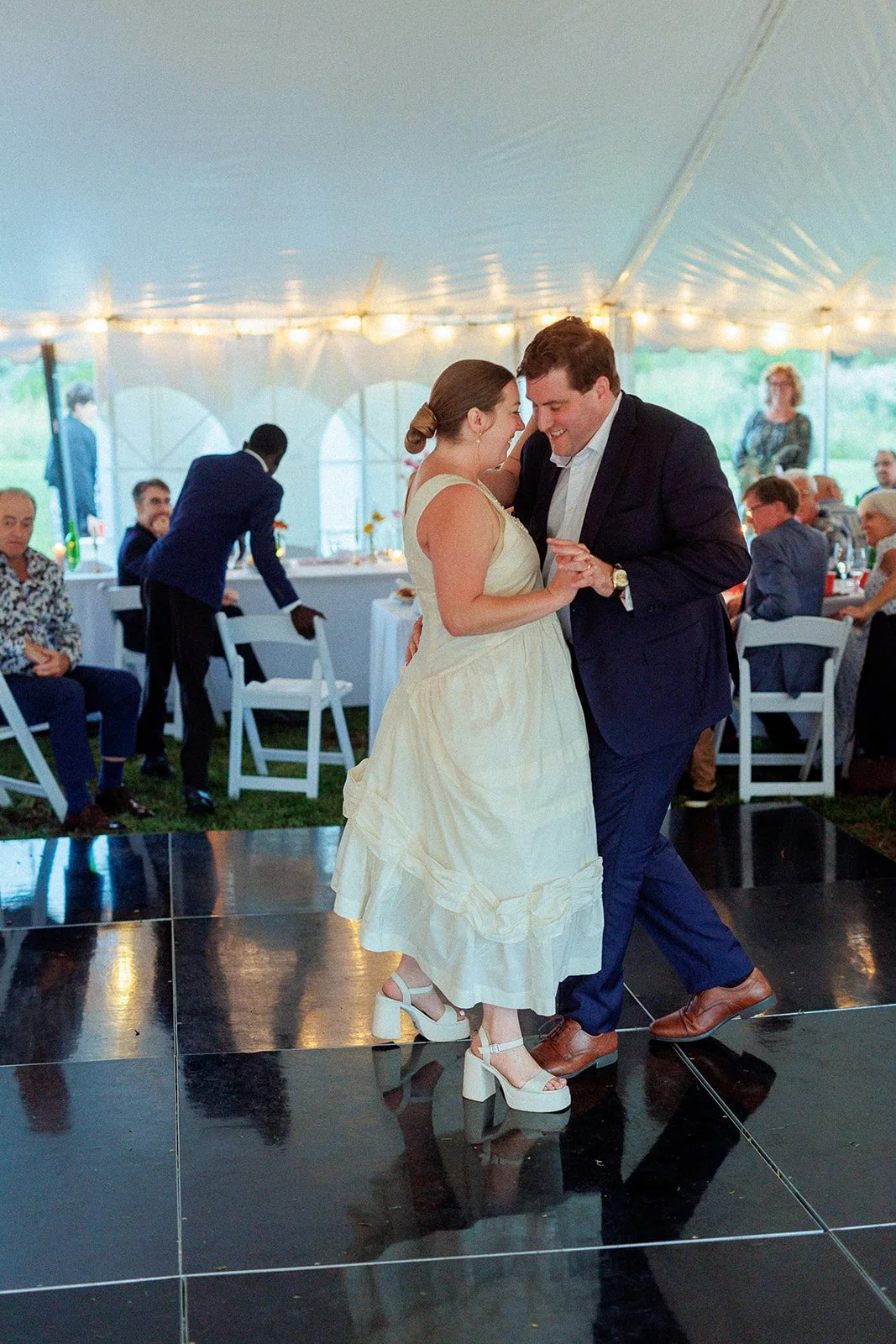 A couple dancing at a wedding reception under a large tent, with guests seated at tables in the background.