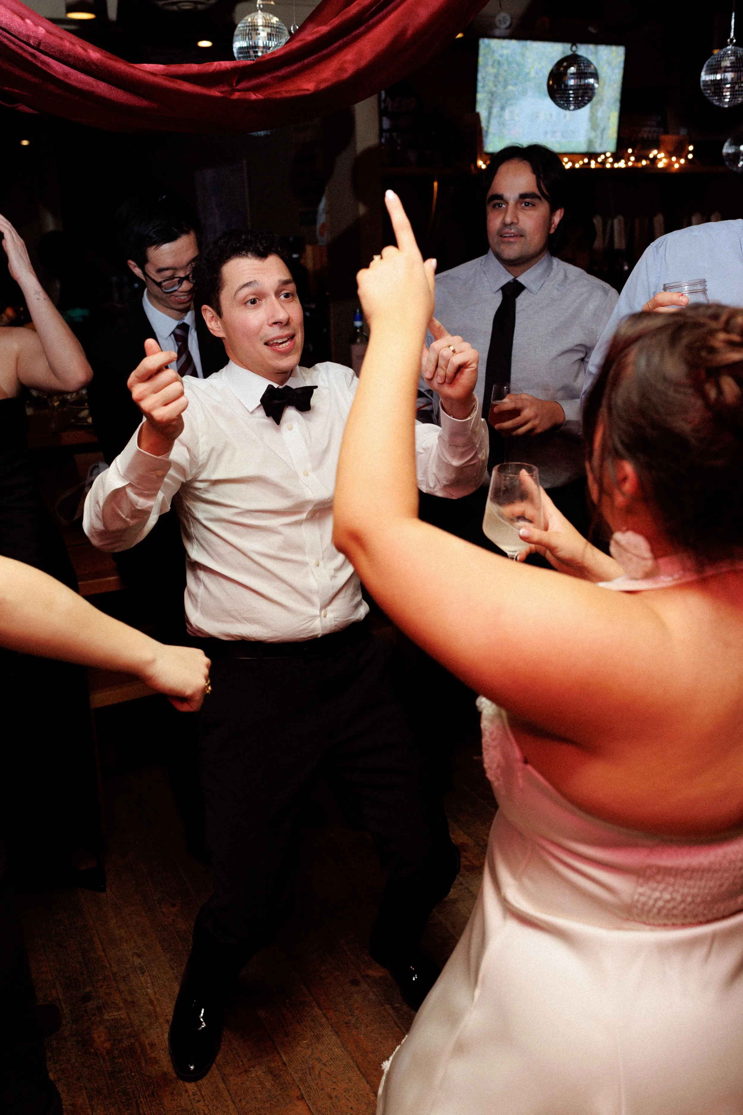People dancing and socializing at a party, with a man in a white shirt and black bowtie dancing in the center.