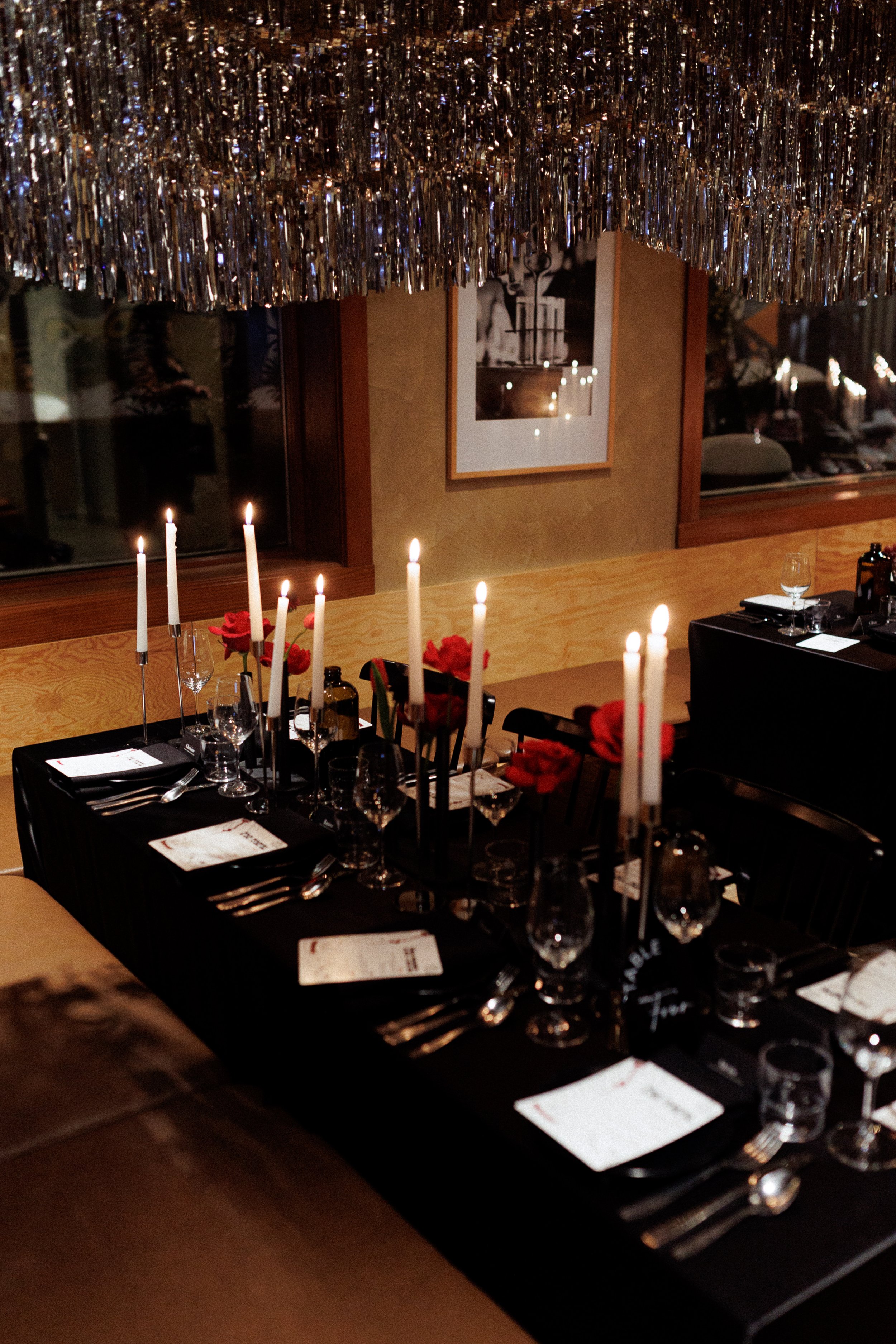 A restaurant table decorated for dinner with tall white candles and red flowers, set with glassware, silverware, and menus, under a shiny hanging decoration.