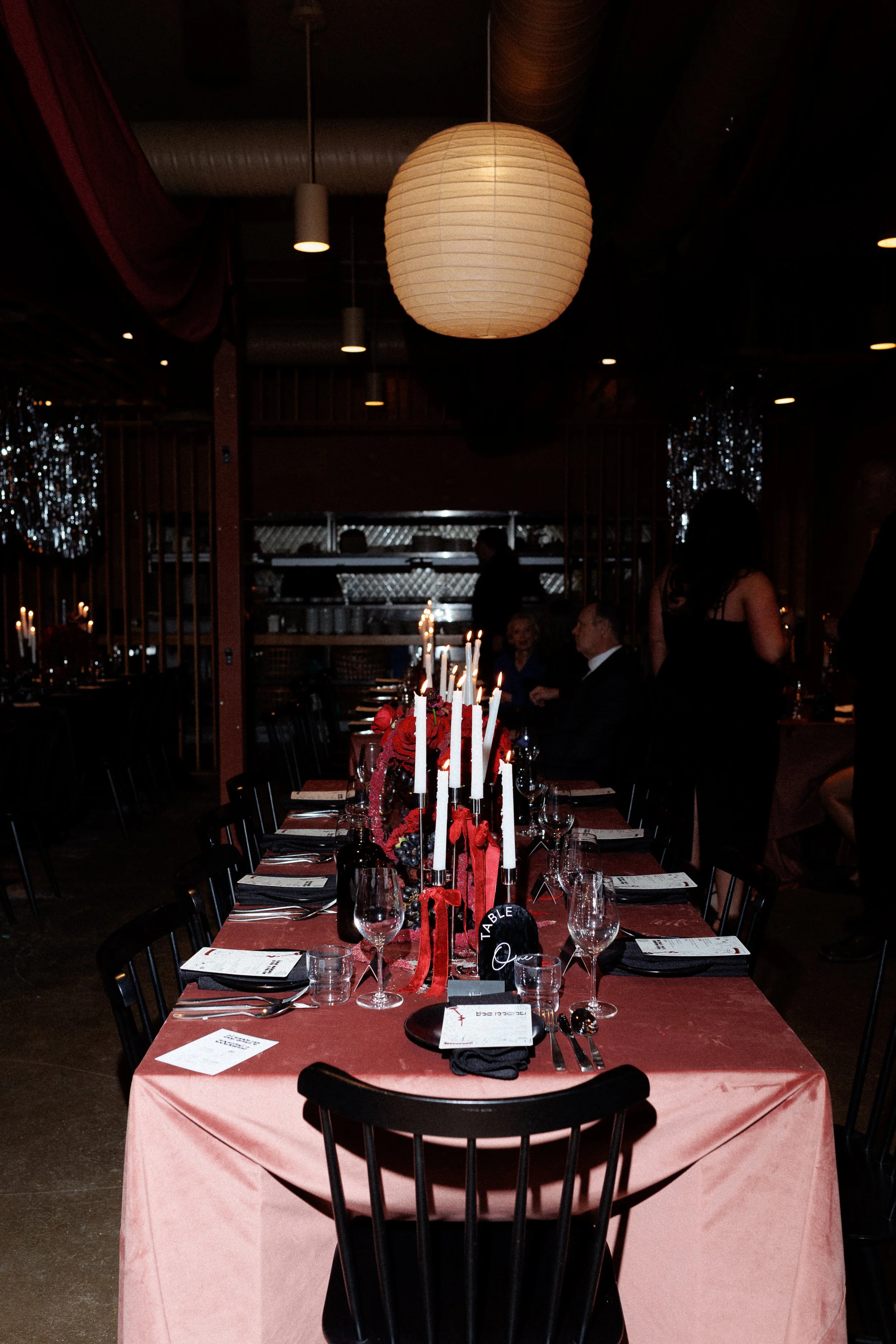 Decorated dining table in a dimly lit restaurant with a pink tablecloth, tall white candles, and floral centerpiece, set for a formal event or dinner.