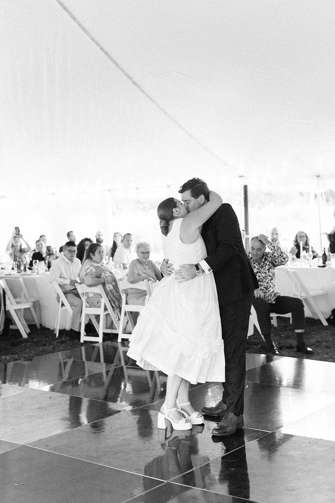A couple sharing a dance at their wedding reception, with guests sitting at tables in the background under a large tent.