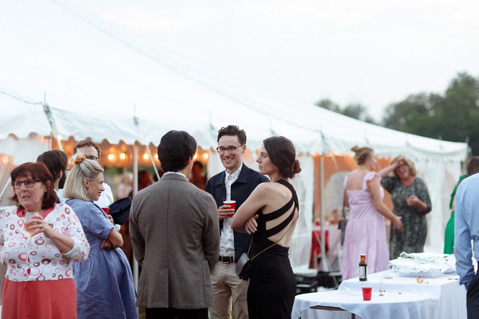 People socializing outdoors at a tented event, some holding drinks, with a table of food and drinks in the foreground.