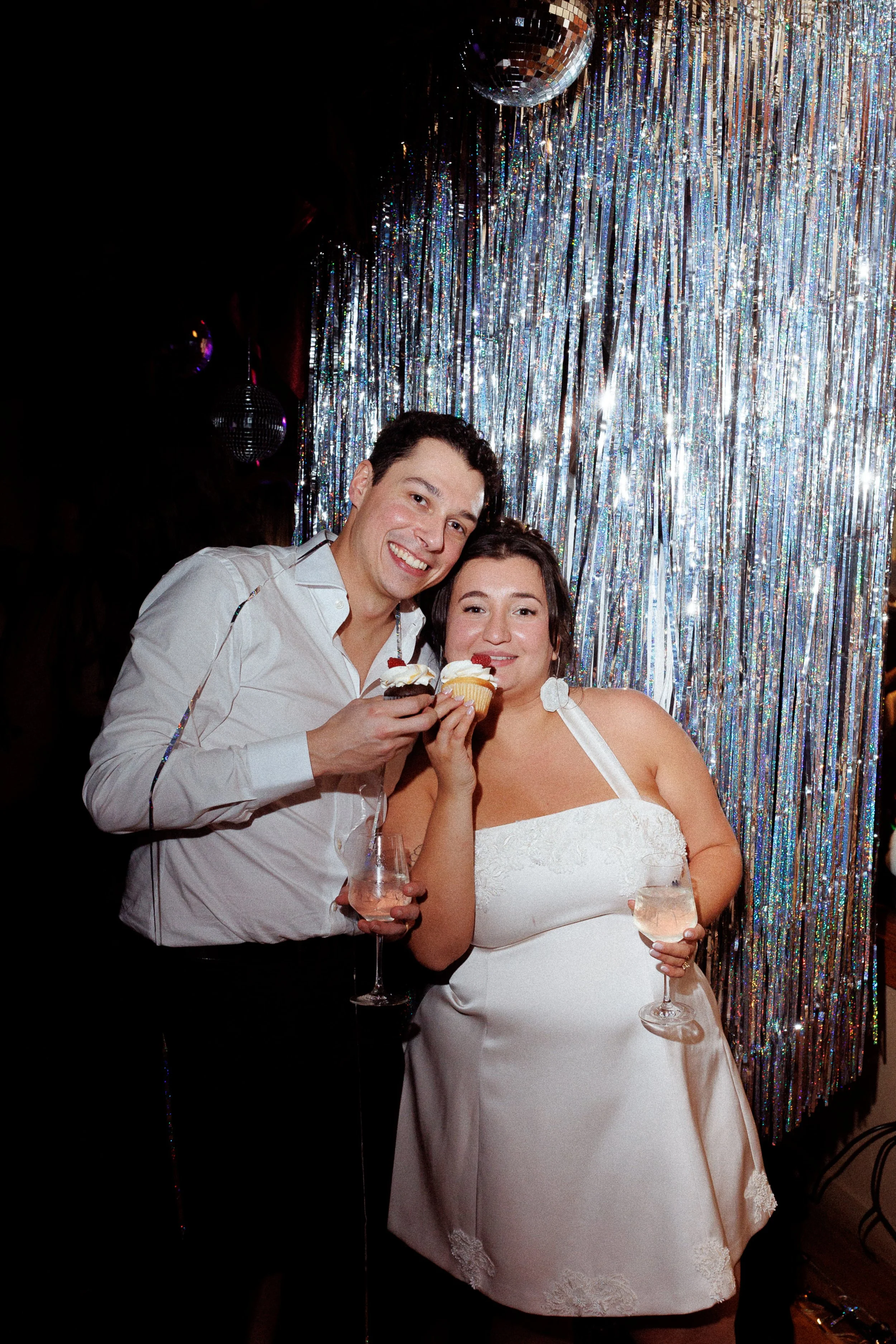 A happy couple in formal attire celebrating at a party, holding cupcakes and wine glasses, against a shiny, metallic, sparkly backdrop with disco balls.