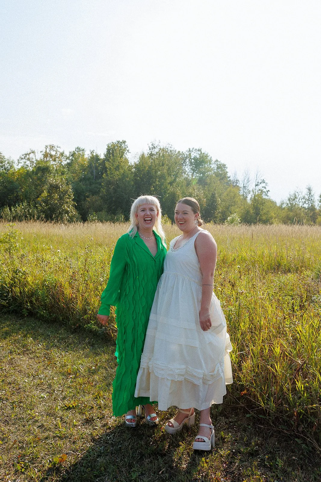 Two women in dressy outfits smiling and standing outside in a grassy field with trees in the background.