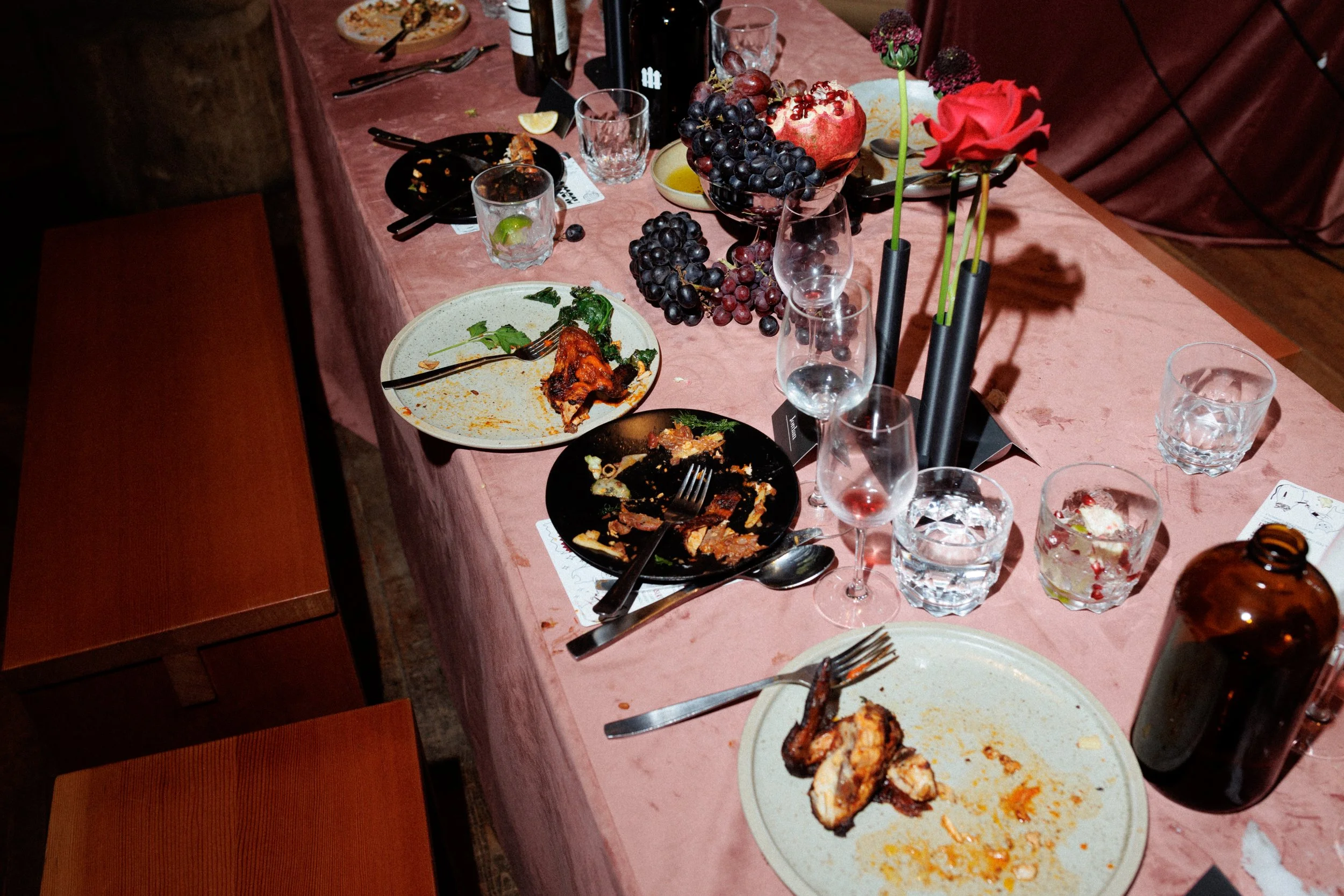 A dining table with empty and partially eaten plates, glasses, and cutlery, along with a centerpiece of grapes and flowers, indicating a recent meal or party.