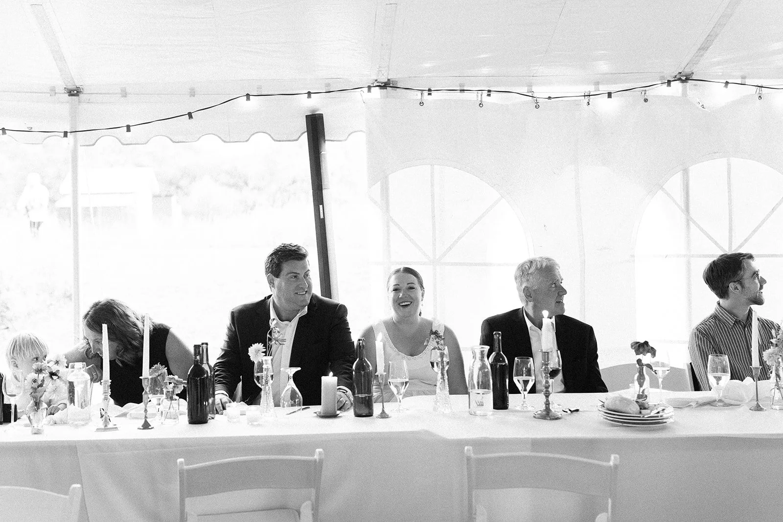 Black and white photo of a wedding reception with six people sitting at a head table decorated with candles, bottles, and flowers. Two young men, a young woman, an older man, and a woman and a young girl are smiling and chatting.