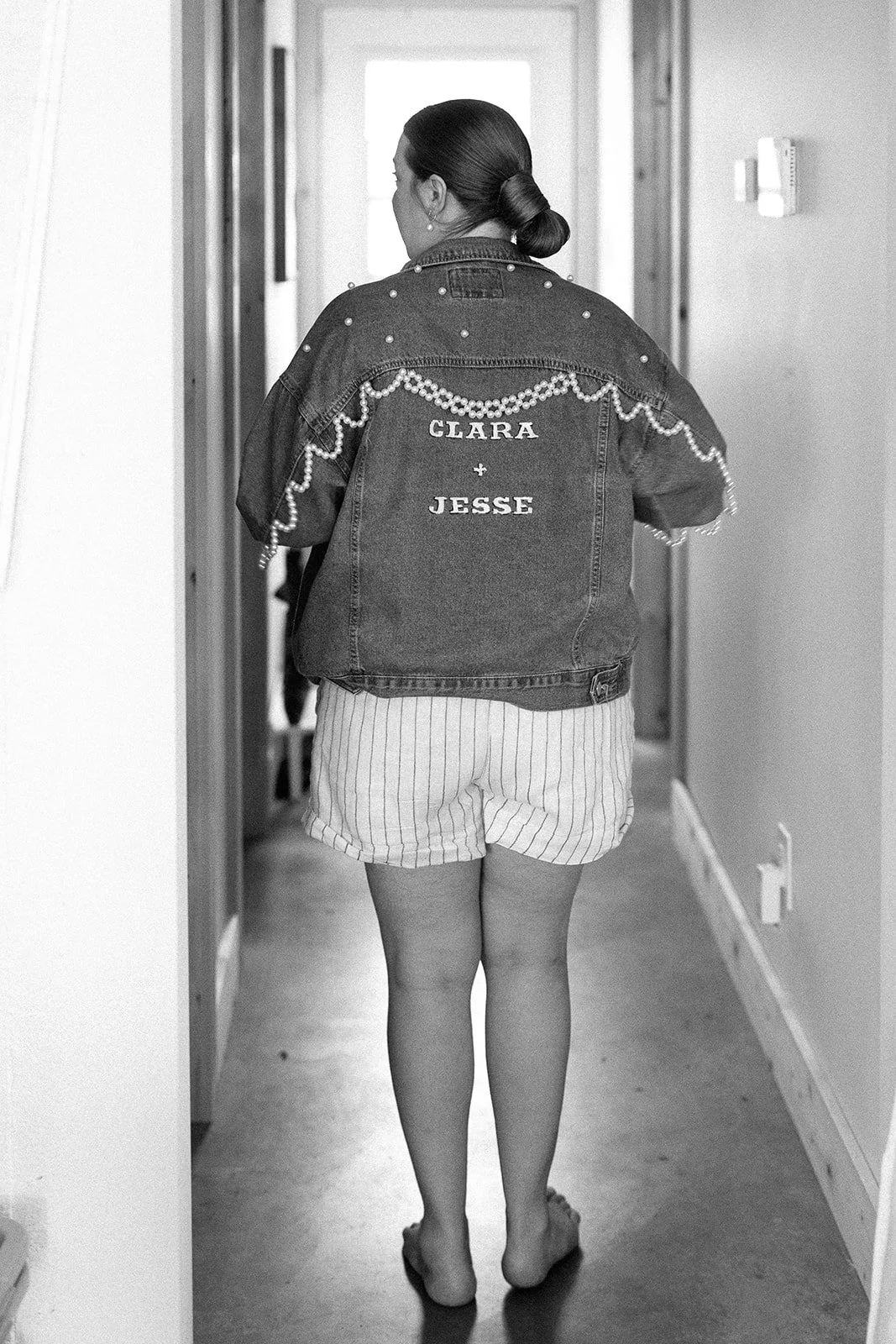 A woman walking down a hallway wearing striped shorts, a denim jacket with pearl decorations and the names 'Clara' and 'Jesse' on the back, and with her hair tied in a low bun.