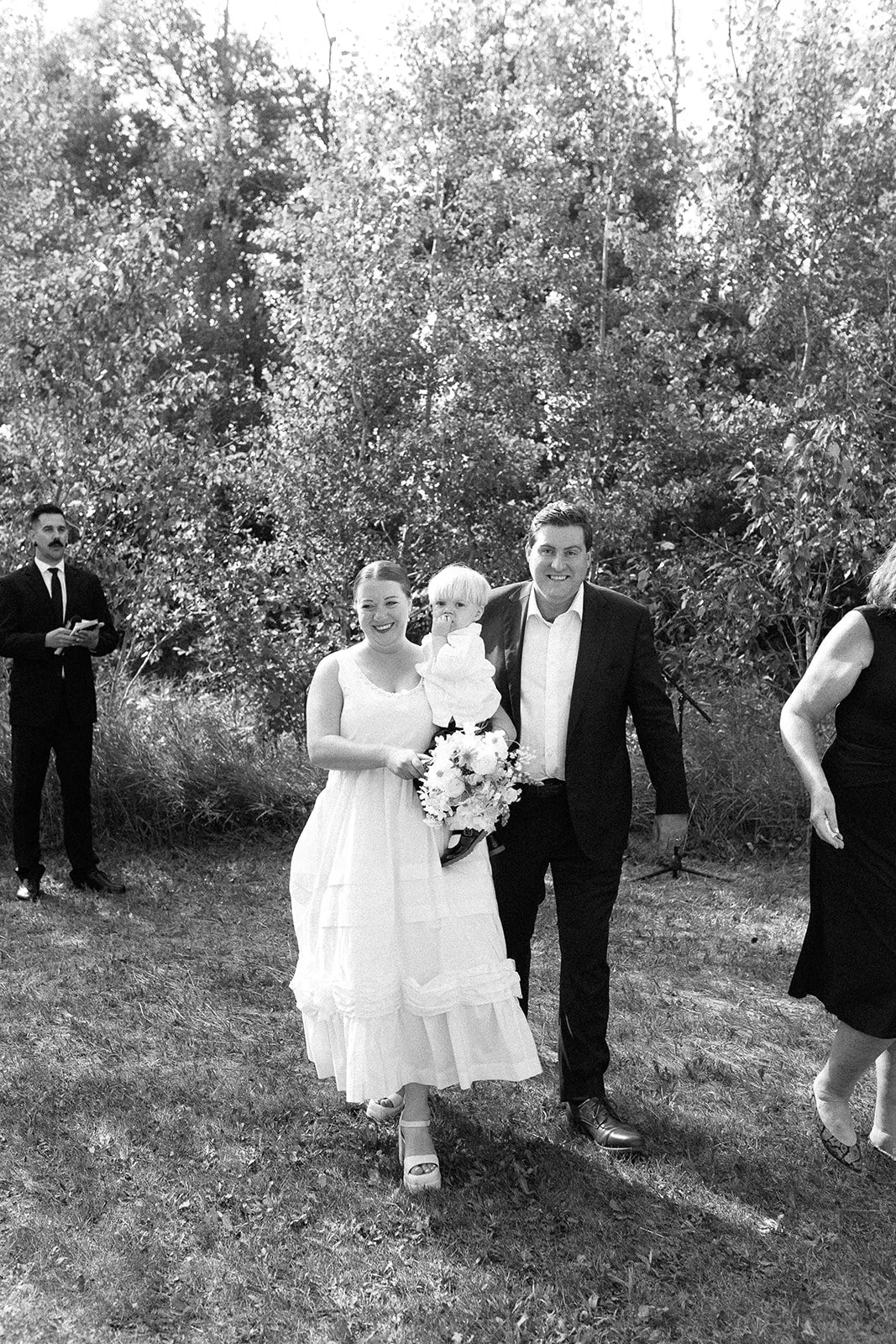 A woman in a white dress, holding a bouquet, walking outdoors with a man in a dark suit and a young child. Other people are seen in the background among trees and bushes.