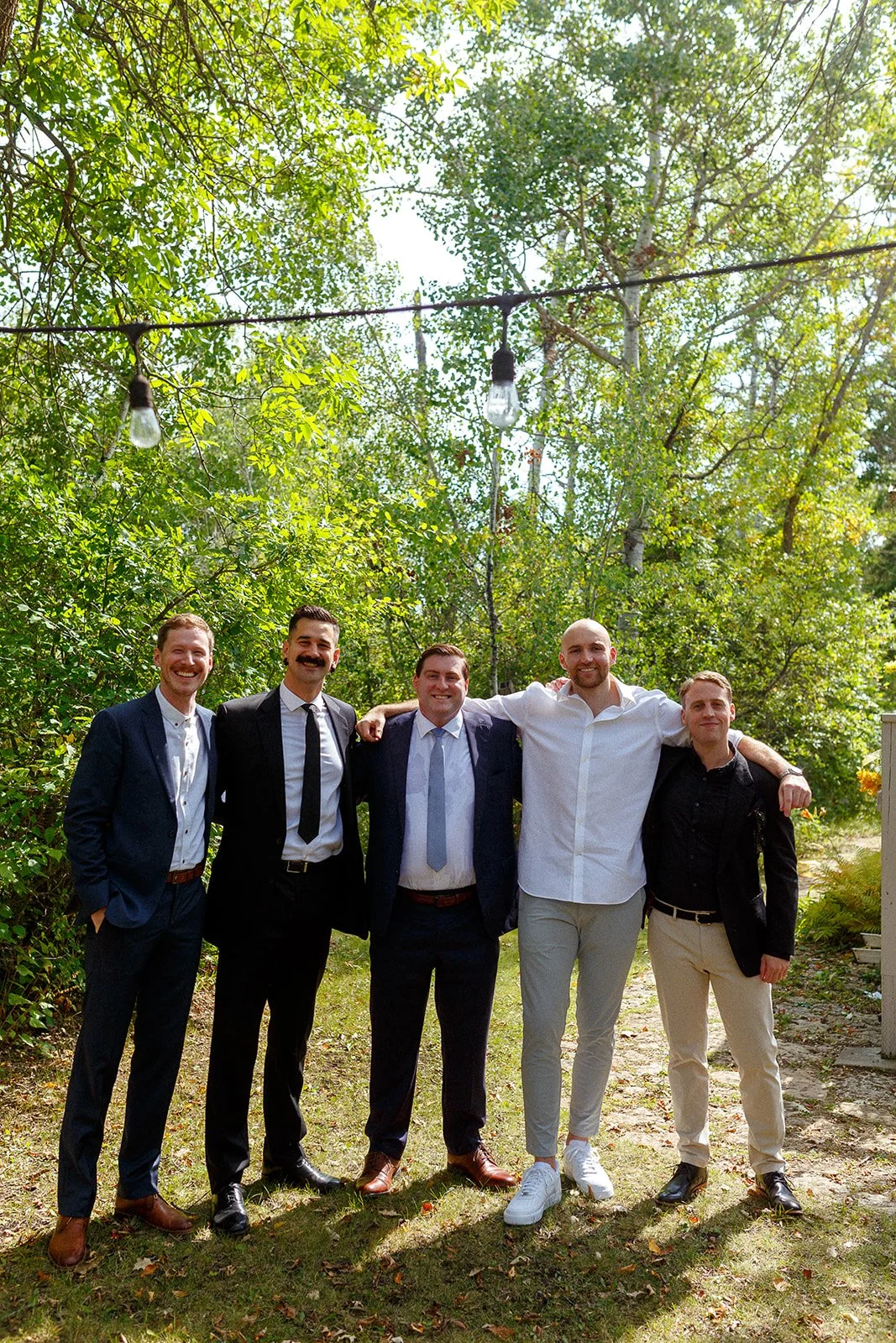 A group of five men in suits standing outdoors in a wooded area, smiling with their arms around each other.