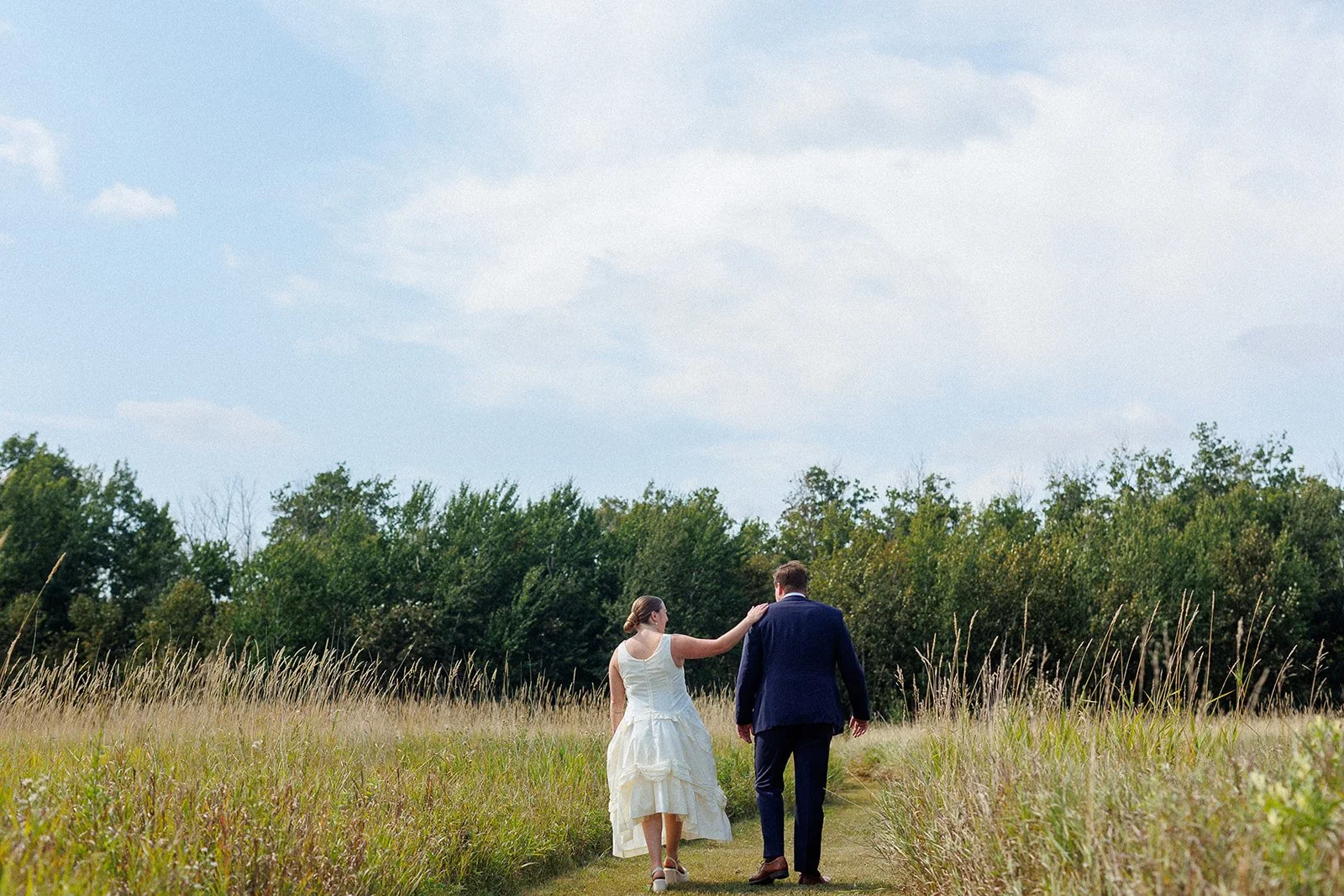 A bride and groom walking hand in hand through a grassy field with trees in the background during the daytime.
