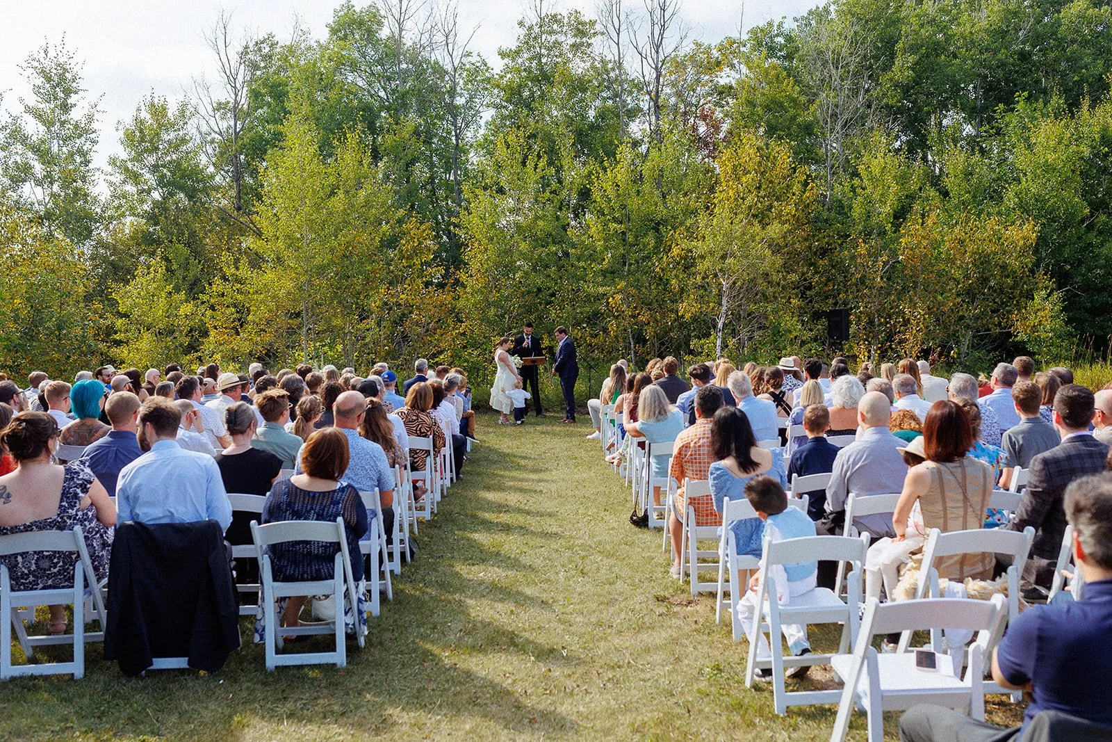 Outdoor wedding ceremony with guests seated in white chairs face a couple exchanging vows, surrounded by green trees.