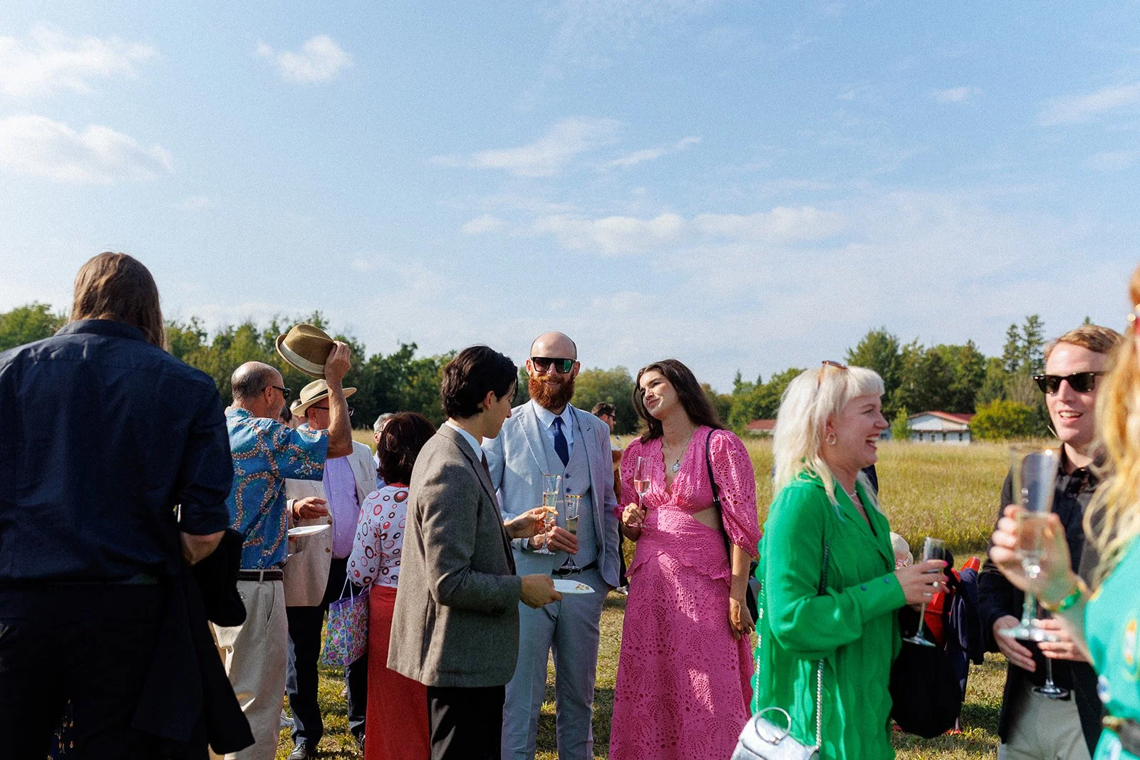 Group of people enjoying an outdoor event, talking and holding drinks in a field on a sunny day.