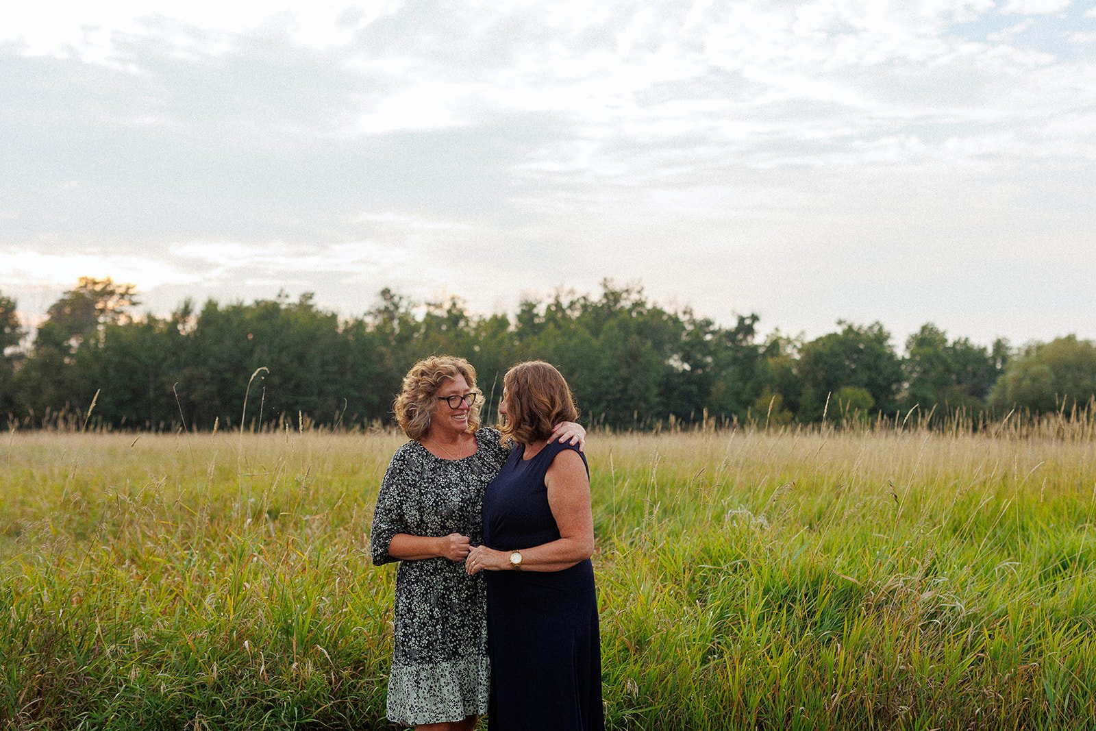 Two women standing in a grassy field, smiling and embracing each other. One woman has curly hair and glasses, wearing a floral dress; the other has straight hair, wearing a sleeveless navy dress. The sky is overcast, and there are trees in the backgr