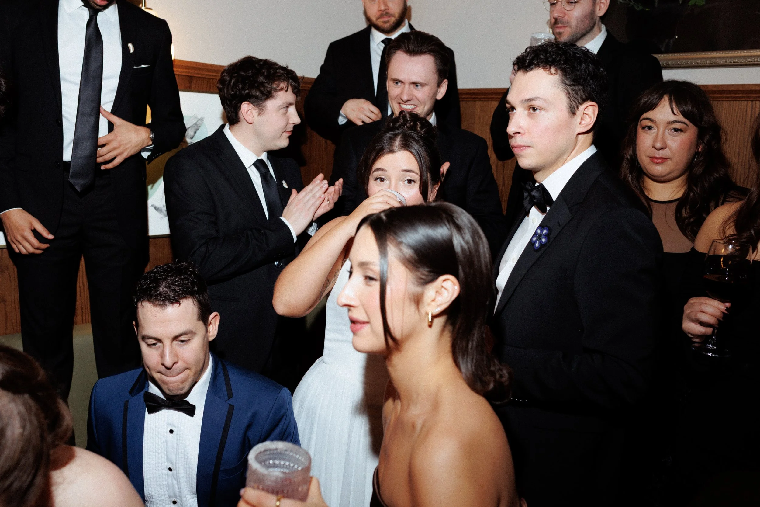 Group of people dressed in formal attire at a social gathering or party, with some holding drinks and engaging in conversations.